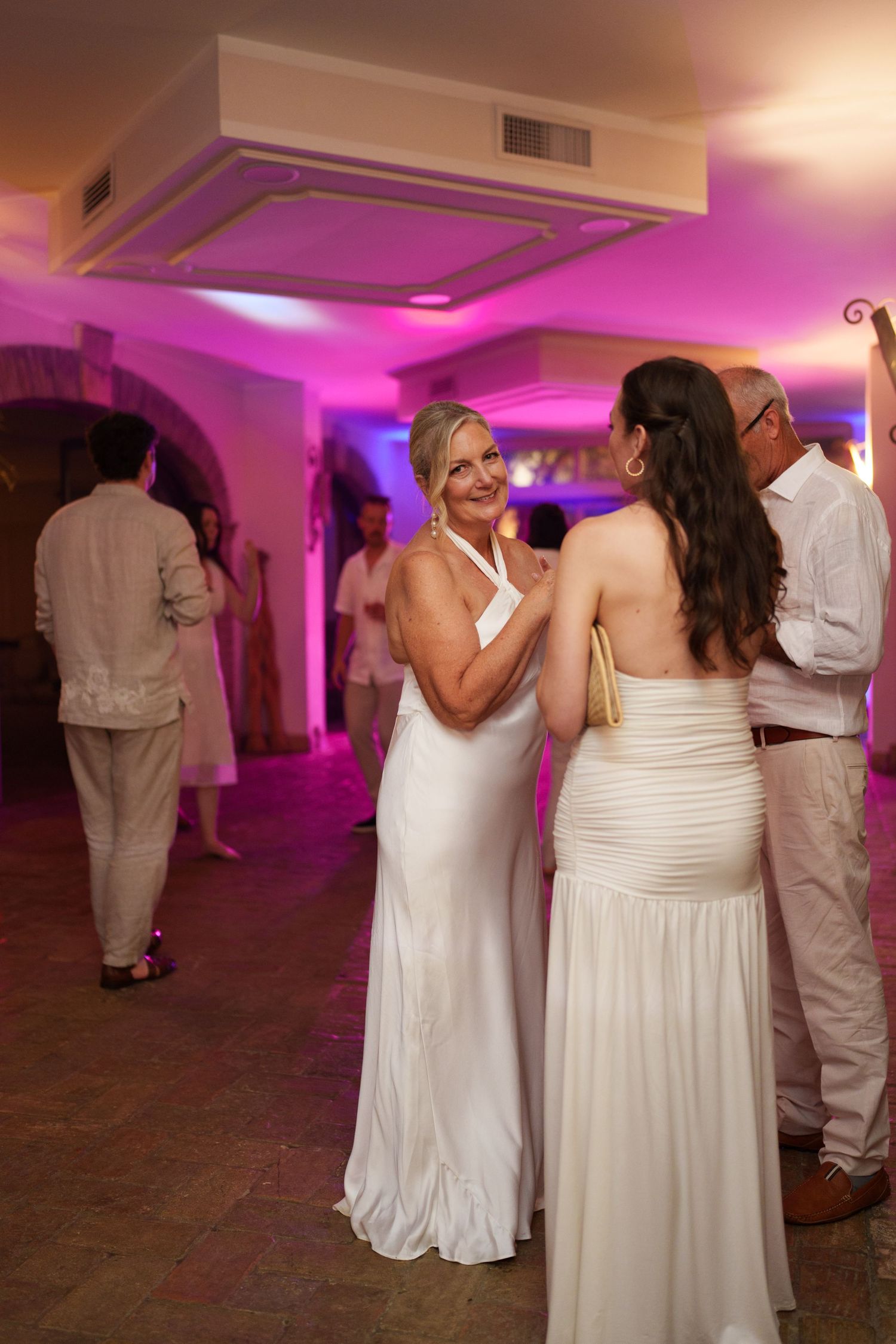 Two women in white gowns dancing at a wedding reception with pink ambient lighting.