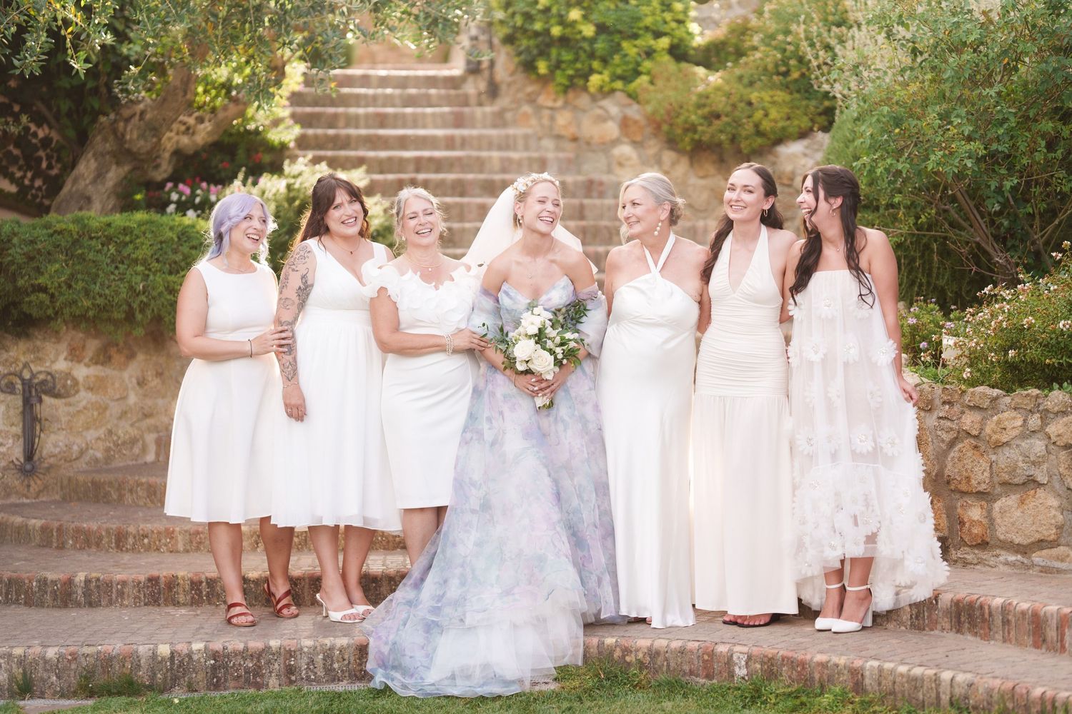 Bride in floral gown poses with six bridesmaids in white dresses outdoors on stone steps surrounded by greenery.