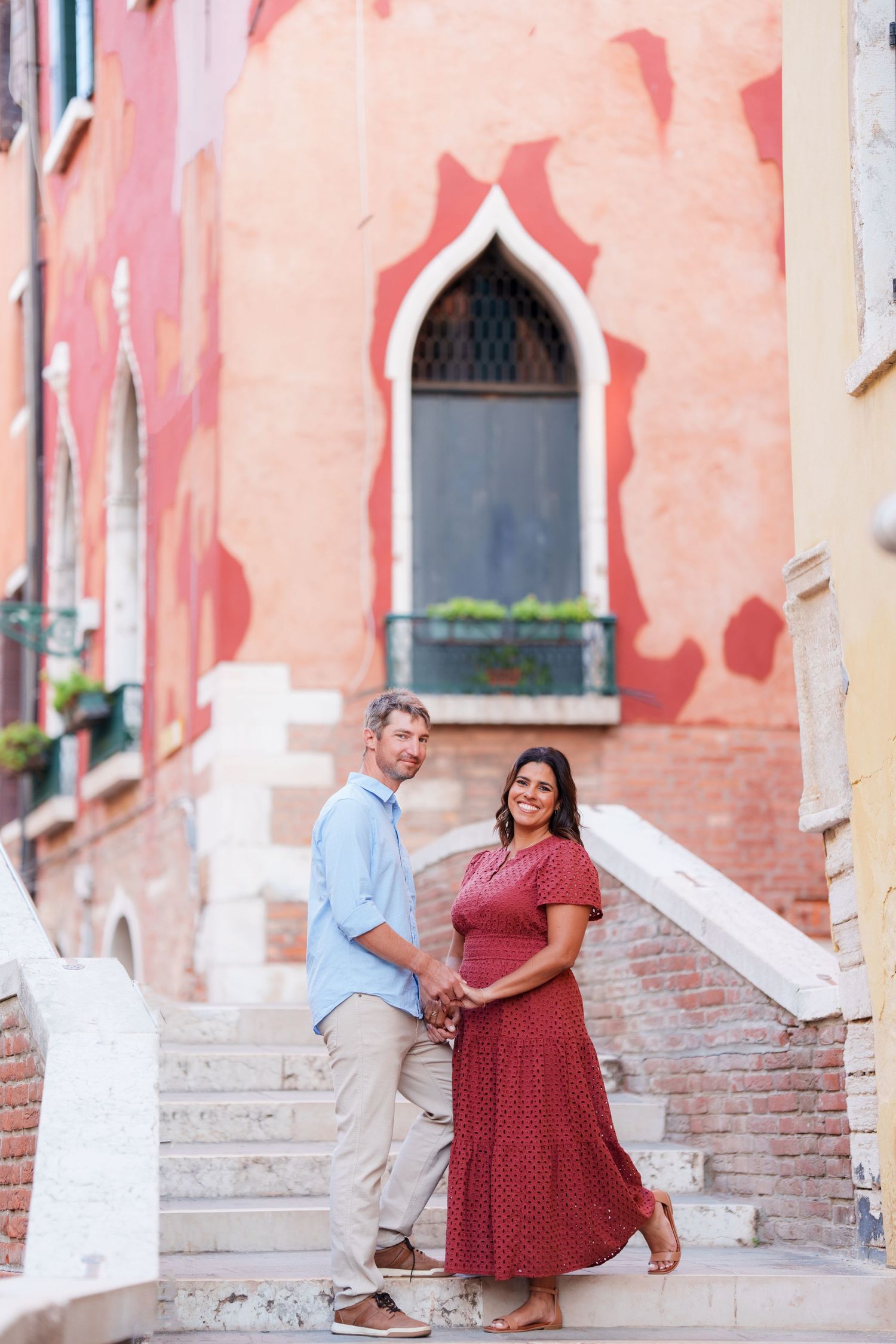 Couple holding hands on stone steps in Venice, Italy, with colorful red and orange historic buildings behind them.