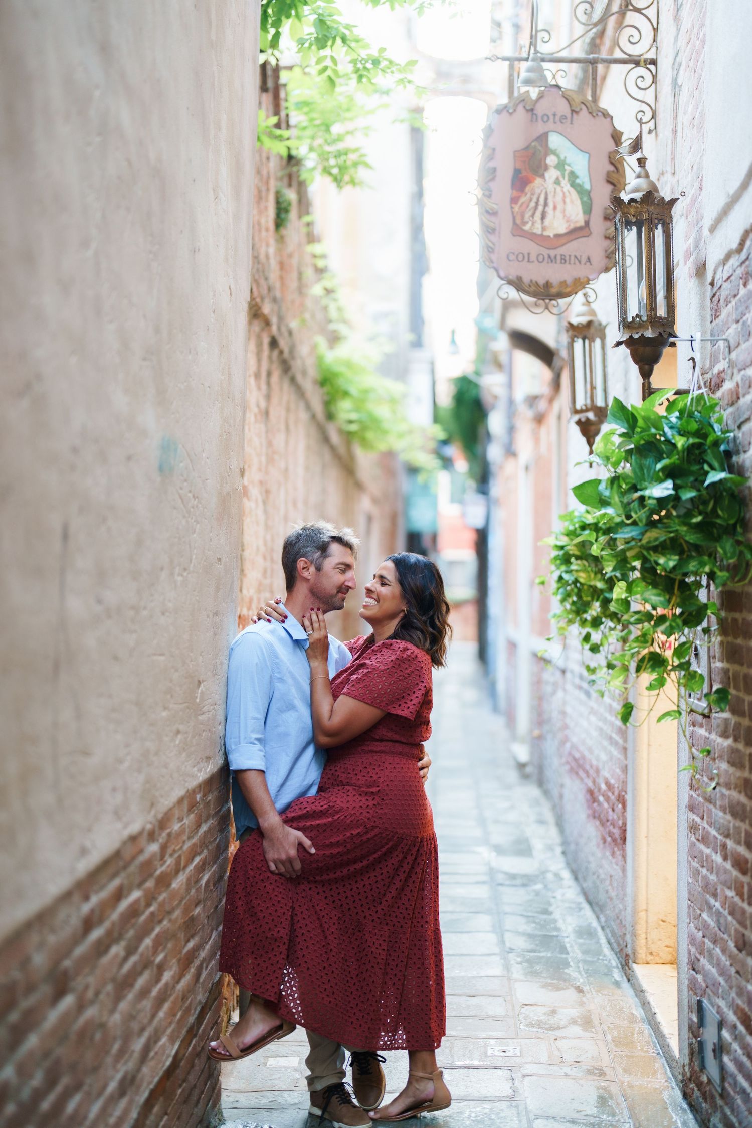 Couple sharing a romantic moment in a narrow European alley, woman in red dress embracing man in blue shirt.