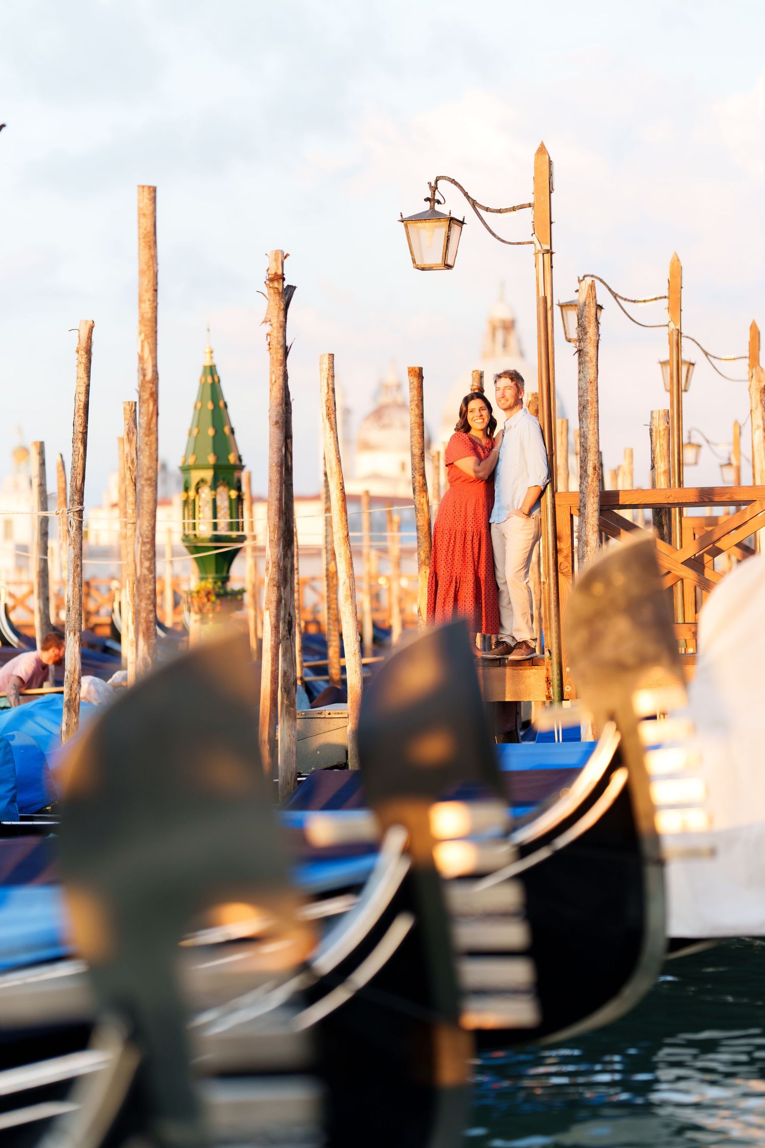 Romantic couple embracing on Venice gondola dock at sunset, with wooden mooring poles and gondolas in foreground.