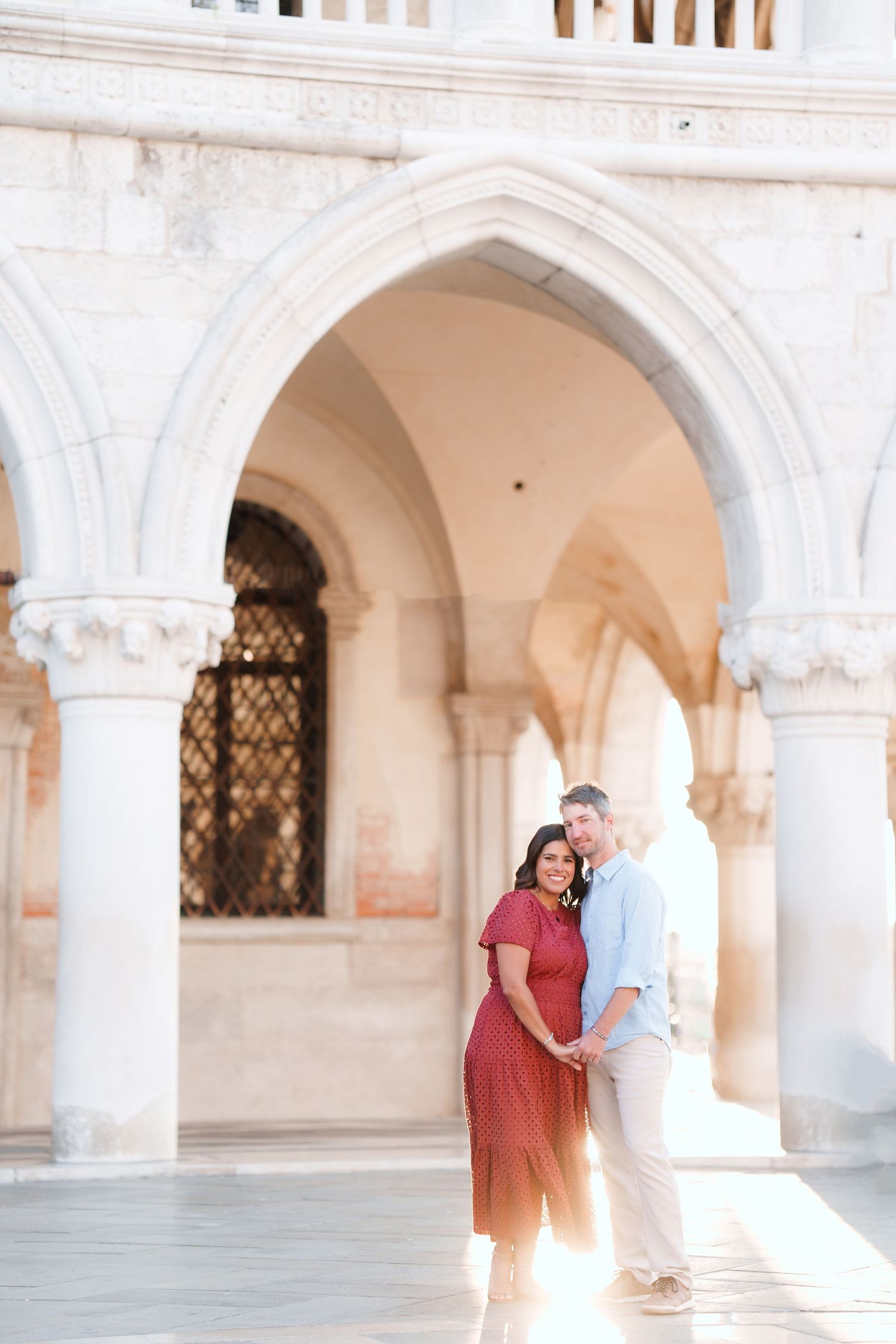 Couple embracing under ornate white marble arches in Venice, woman in red dress, man in light blue shirt, romantic setting.