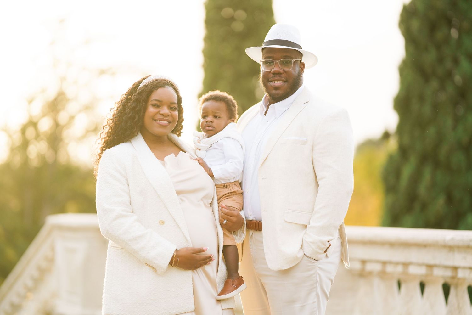 A joyful family in white outfits poses outdoors, with a pregnant woman, a man in a hat, and their toddler son.