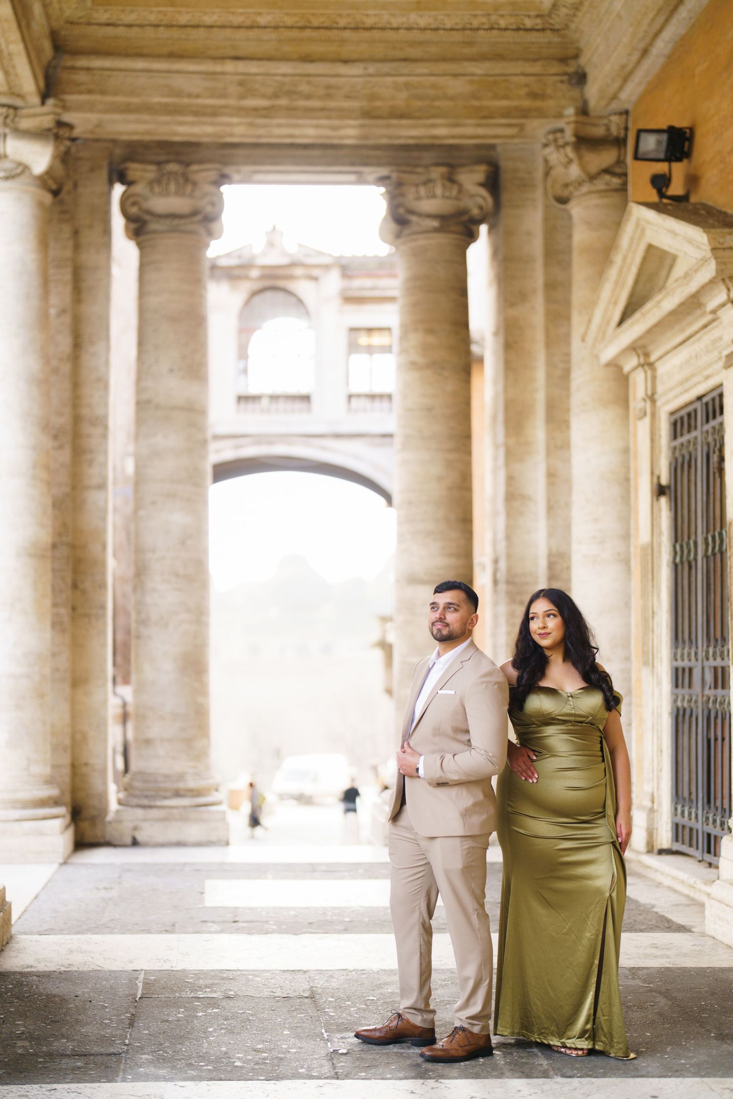 Couple posing elegantly among grand stone columns, woman in olive satin gown, man in beige suit, classical architecture backdrop.