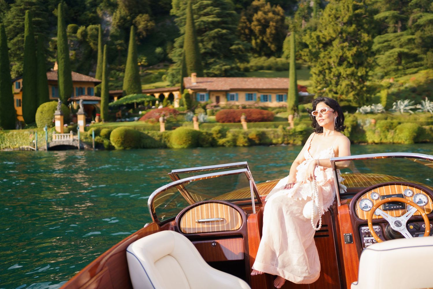 Elegant woman in a white dress sitting on a classic wooden speedboat on a serene lake with lush greenery.