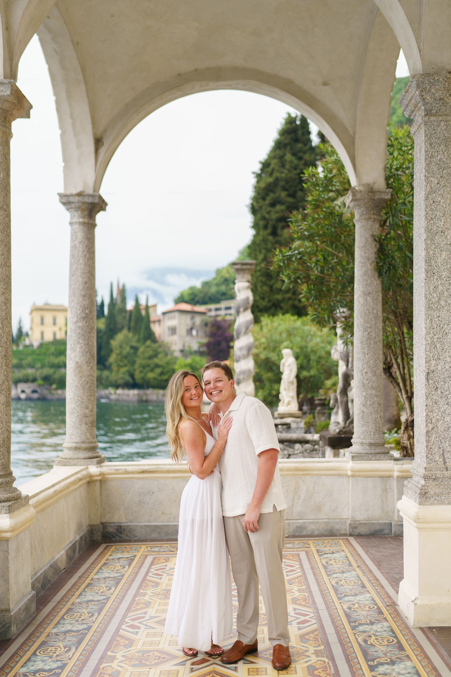 Couple embracing under elegant stone arches at Villa Monastero on Lake Como, Italy, surrounded by statues and scenic water views.