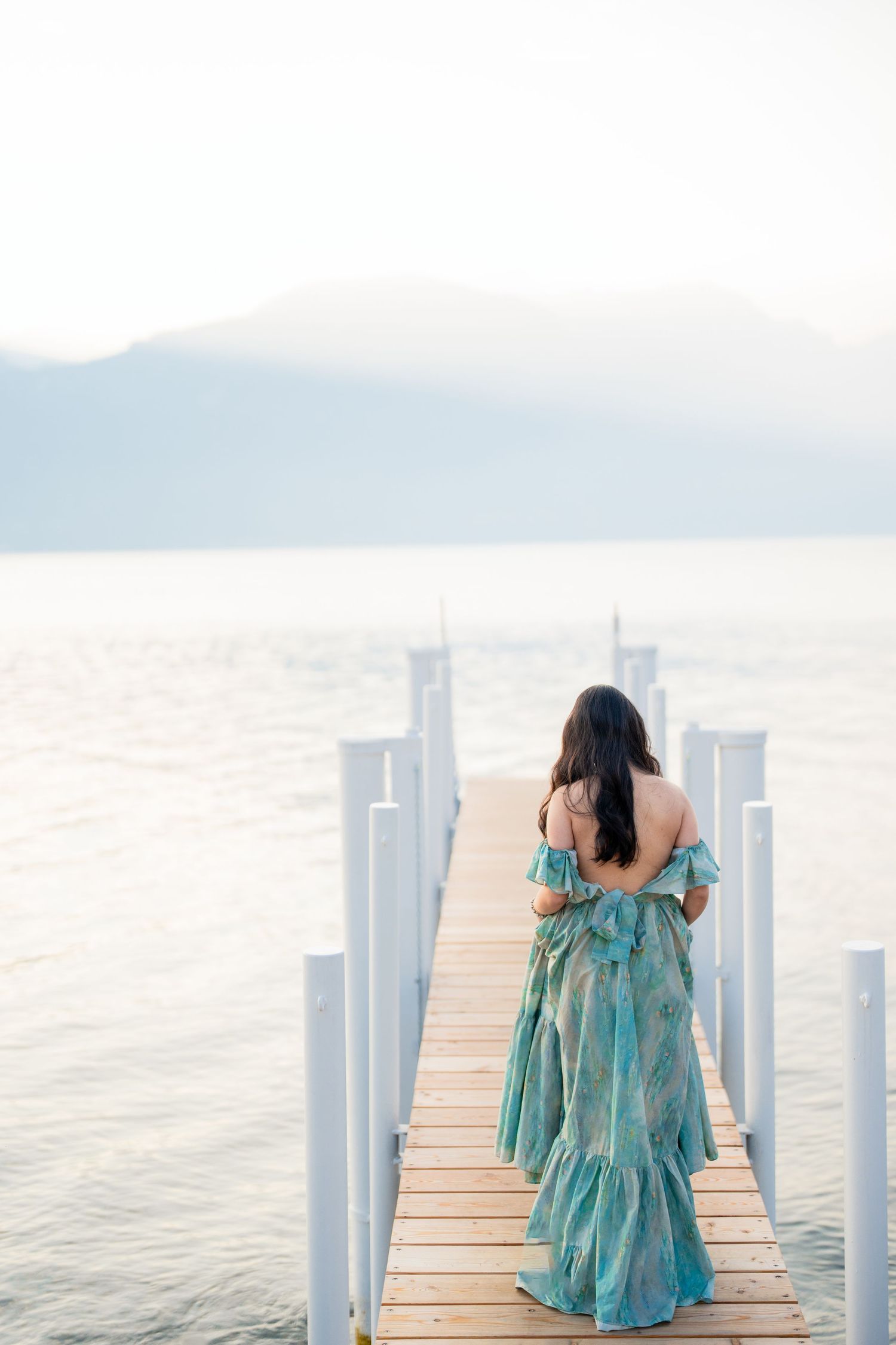 Woman in teal floral off-shoulder gown walks along wooden pier overlooking calm lake with misty mountains.
