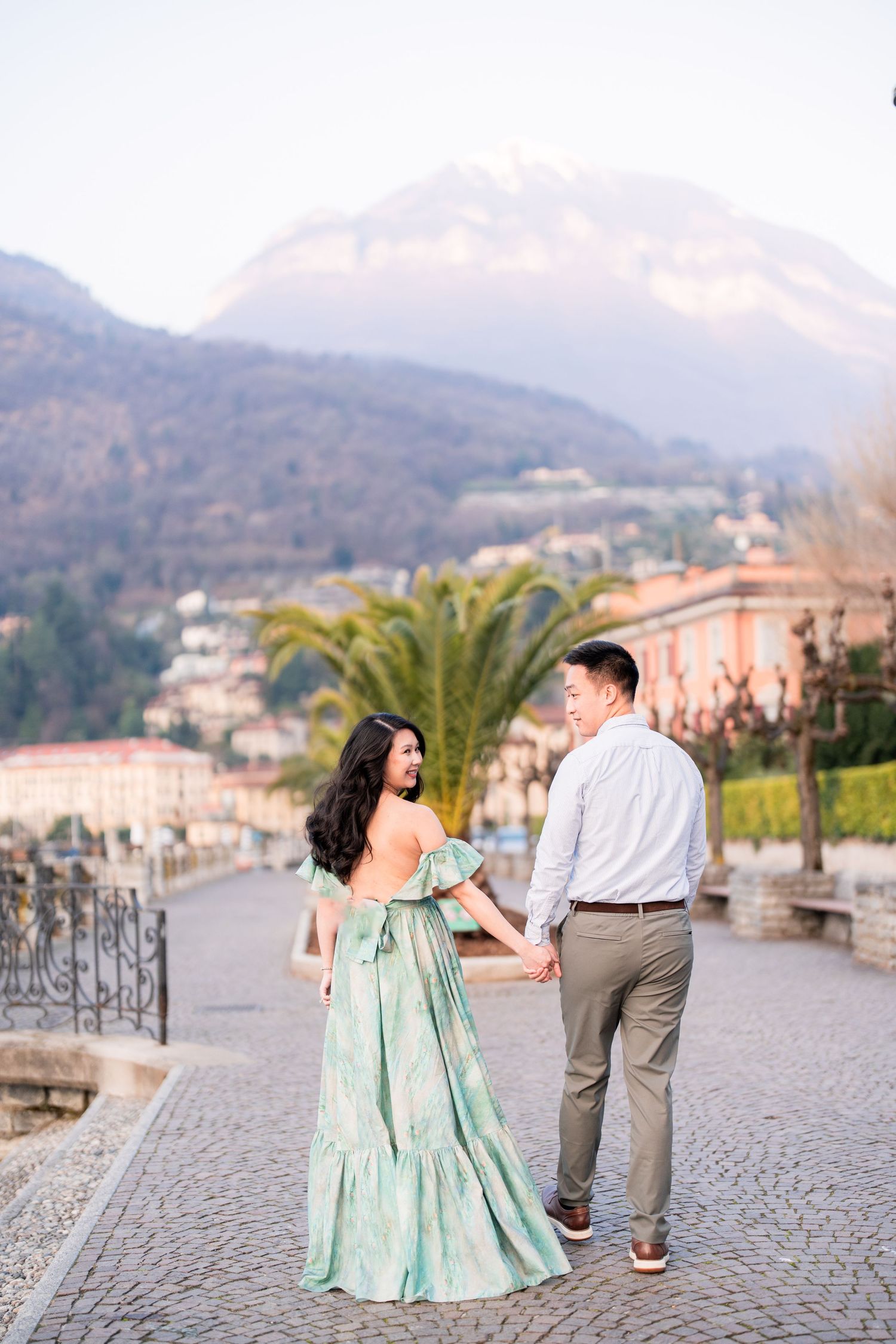 Couple holding hands on cobblestone path with mountain backdrop and palm trees in elegant resort town setting.