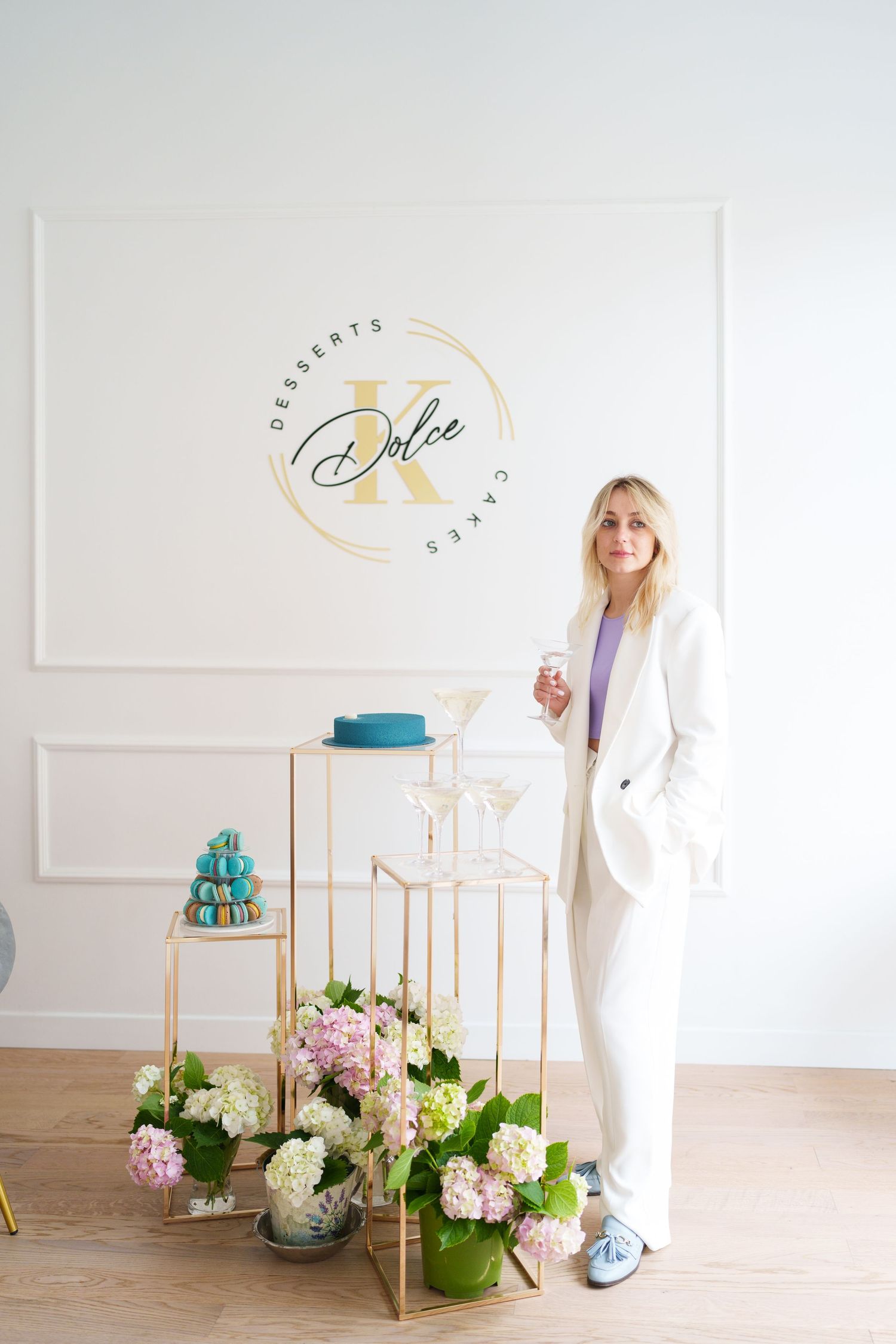 Woman in white suit poses at Dolce Desserts & Cakes shop with macarons, flowers, and champagne glasses.