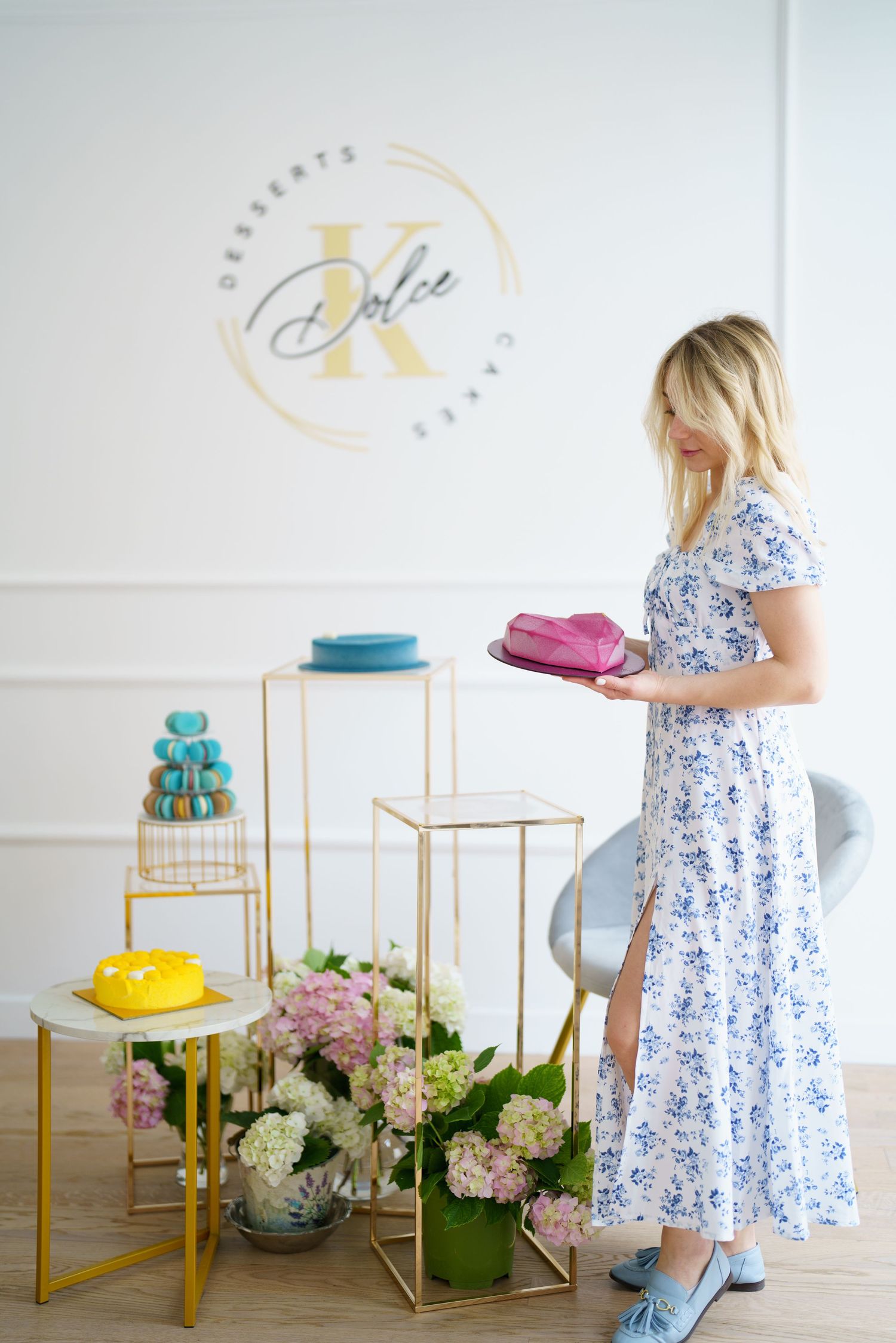 Woman in floral dress holding pink cake at Dolce K desserts shop with colorful cakes and hydrangeas on display.