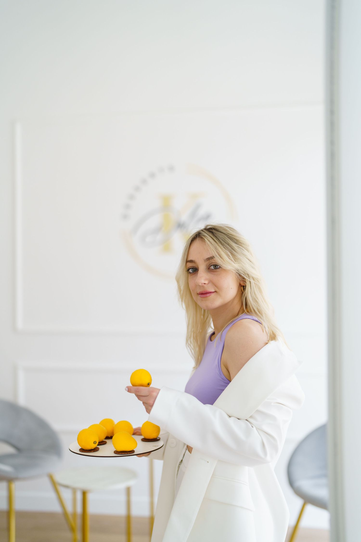 Blonde woman in white blazer and purple top holding yellow macarons on a tray in a bright modern interior.