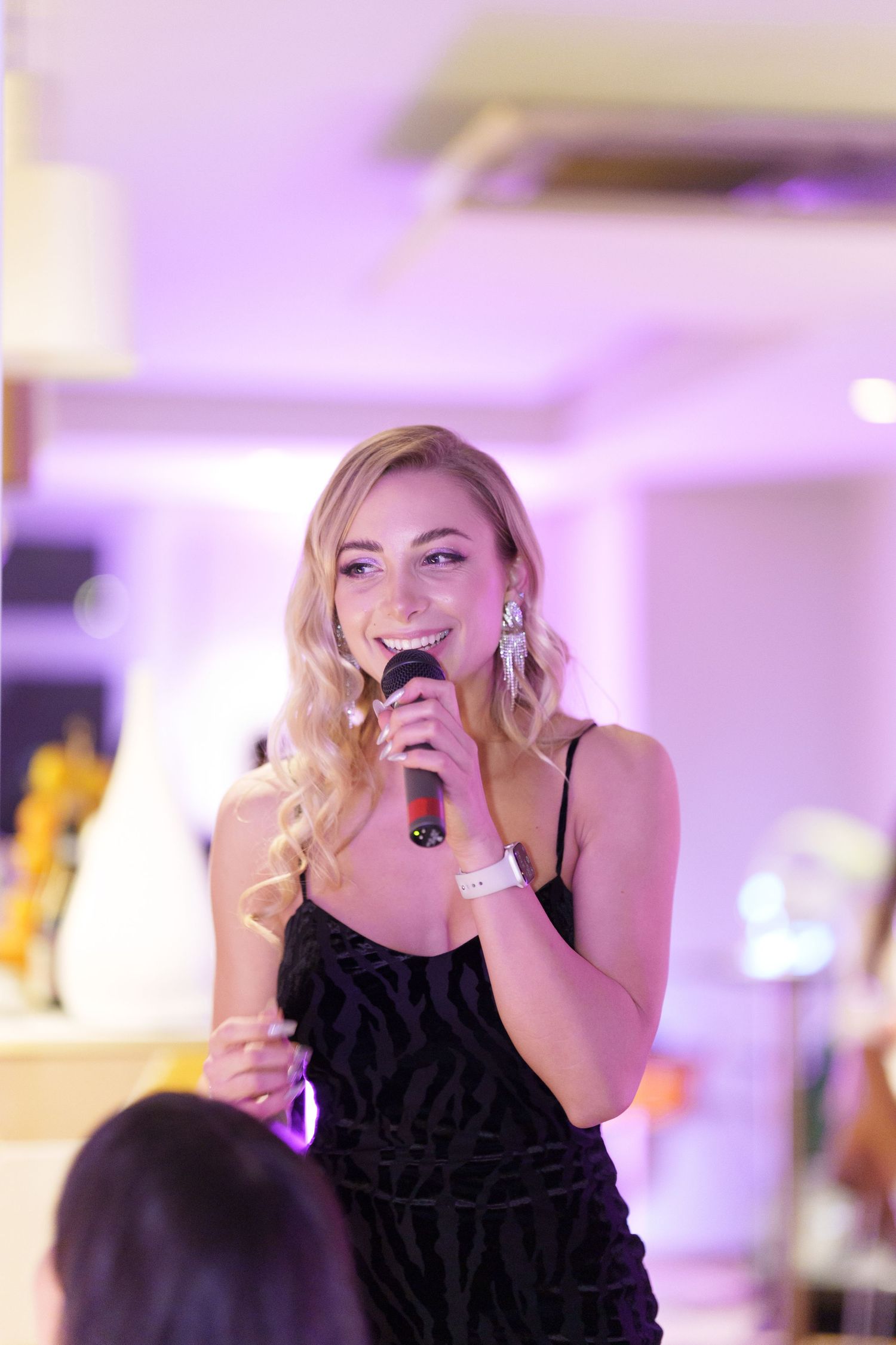 Smiling blonde woman in black dress singing into microphone at elegant event with purple ambient lighting.