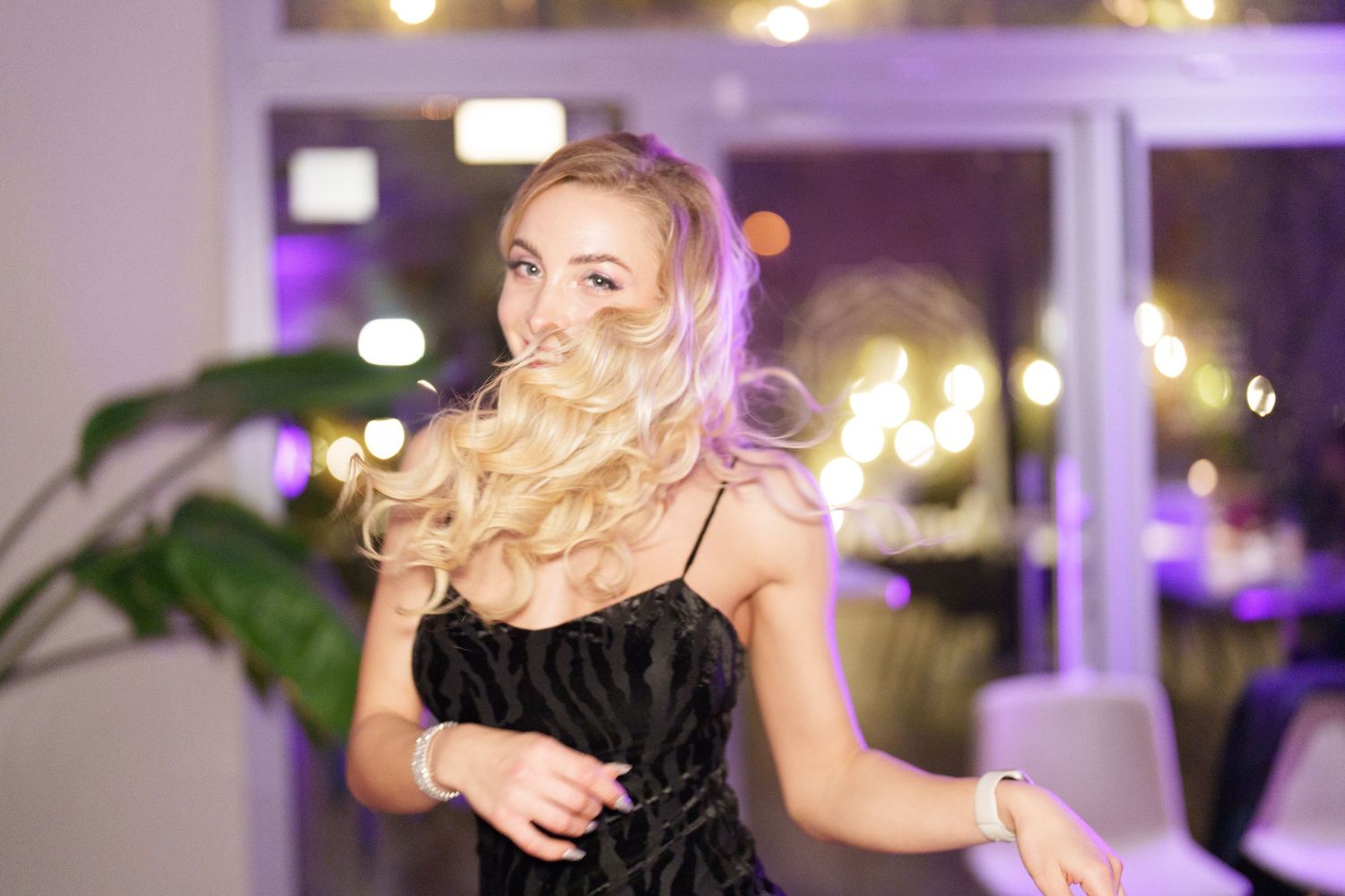 Blonde woman in black dress with curly hair flowing, posing at an elegant venue with bokeh lights.