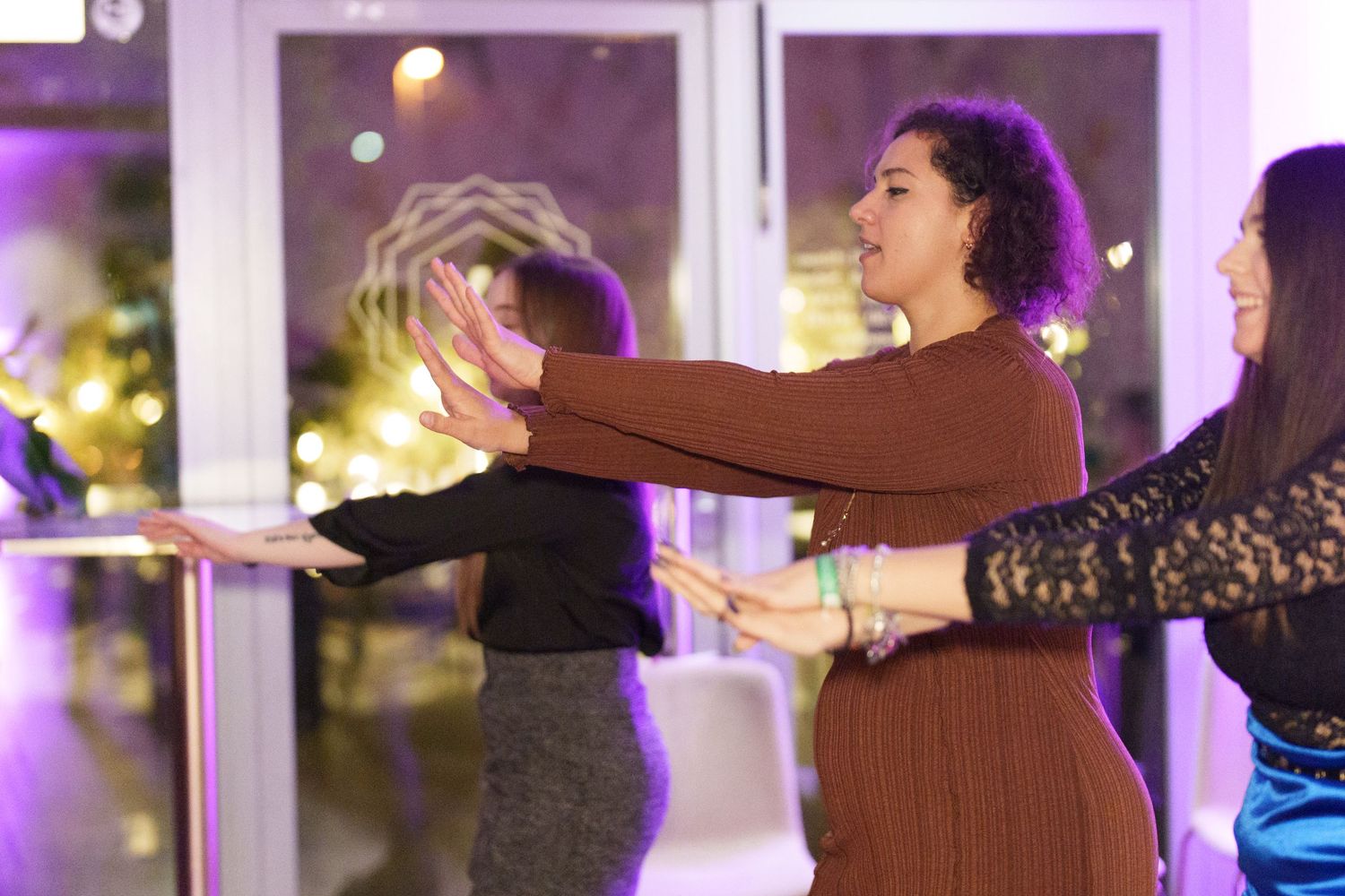 Women dancing together in a line at an evening event, stretching arms forward in a festive indoor venue.