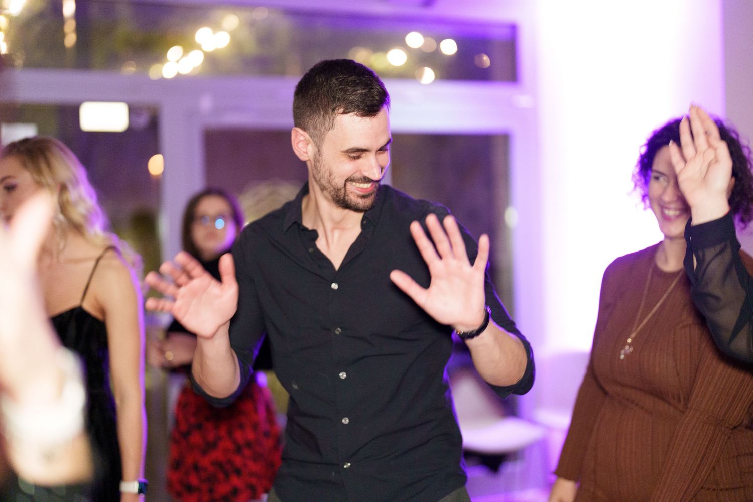 Man in black shirt dancing energetically at a party with other guests and purple ambient lighting.