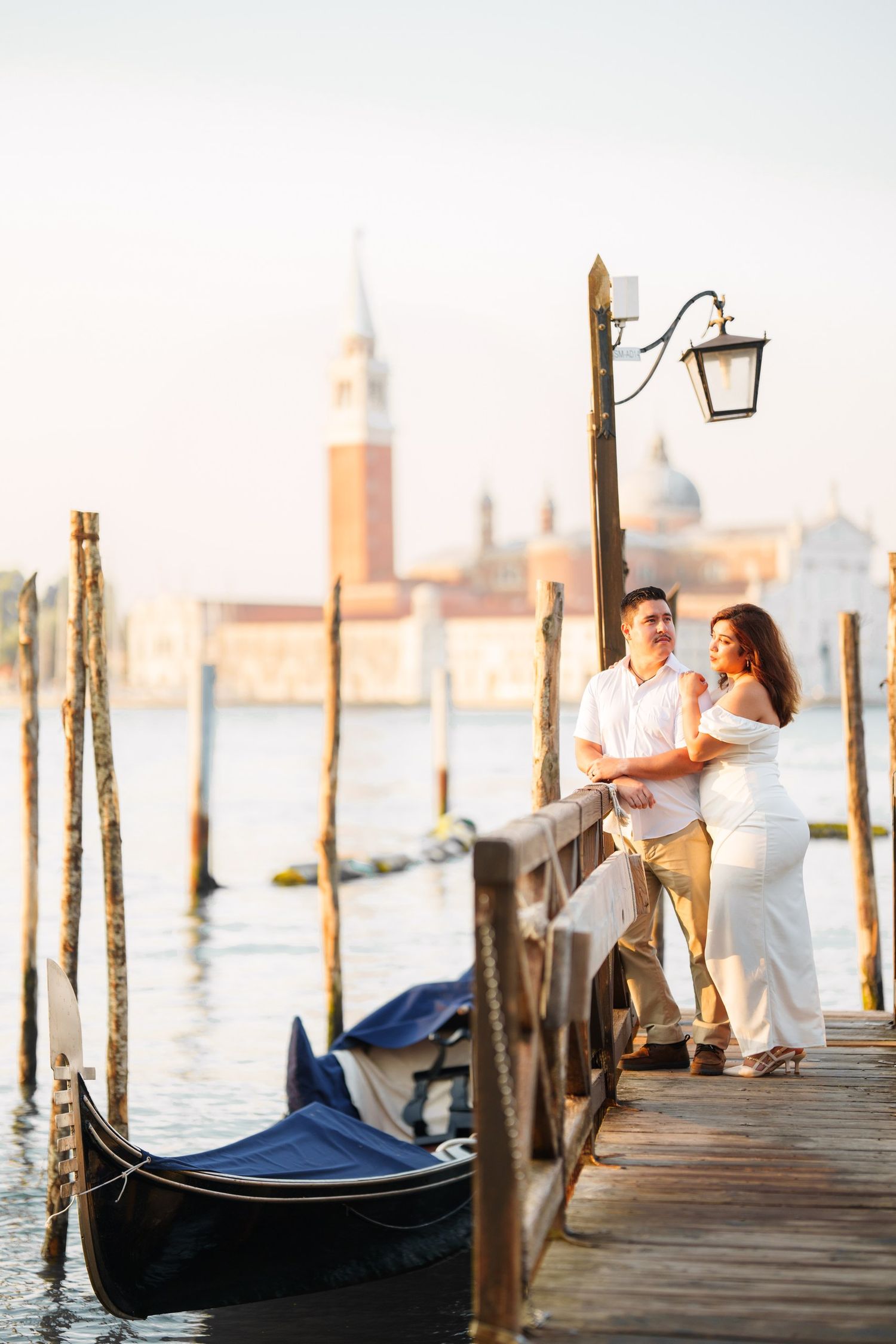 Couple in white outfits on a Venice gondola dock with San Giorgio Maggiore church in the background.