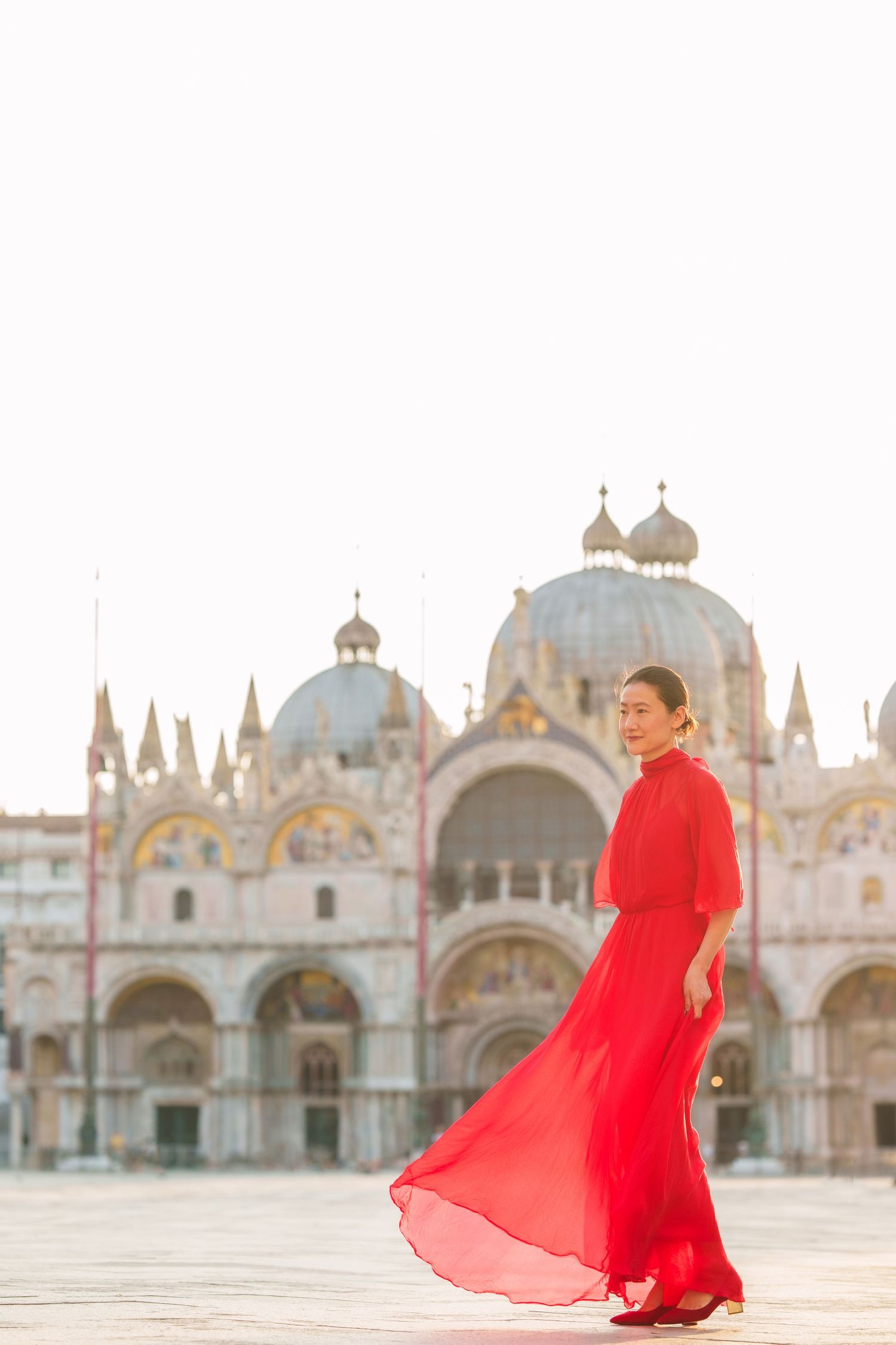 Woman in flowing red dress poses before St. Mark's Basilica in Venice's iconic Piazza San Marco.