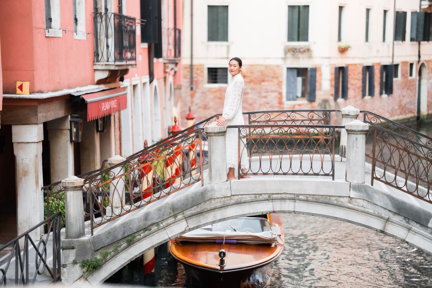 A woman in white dress stands on ornate bridge over Venice canal with moored wooden boat and colorful buildings.