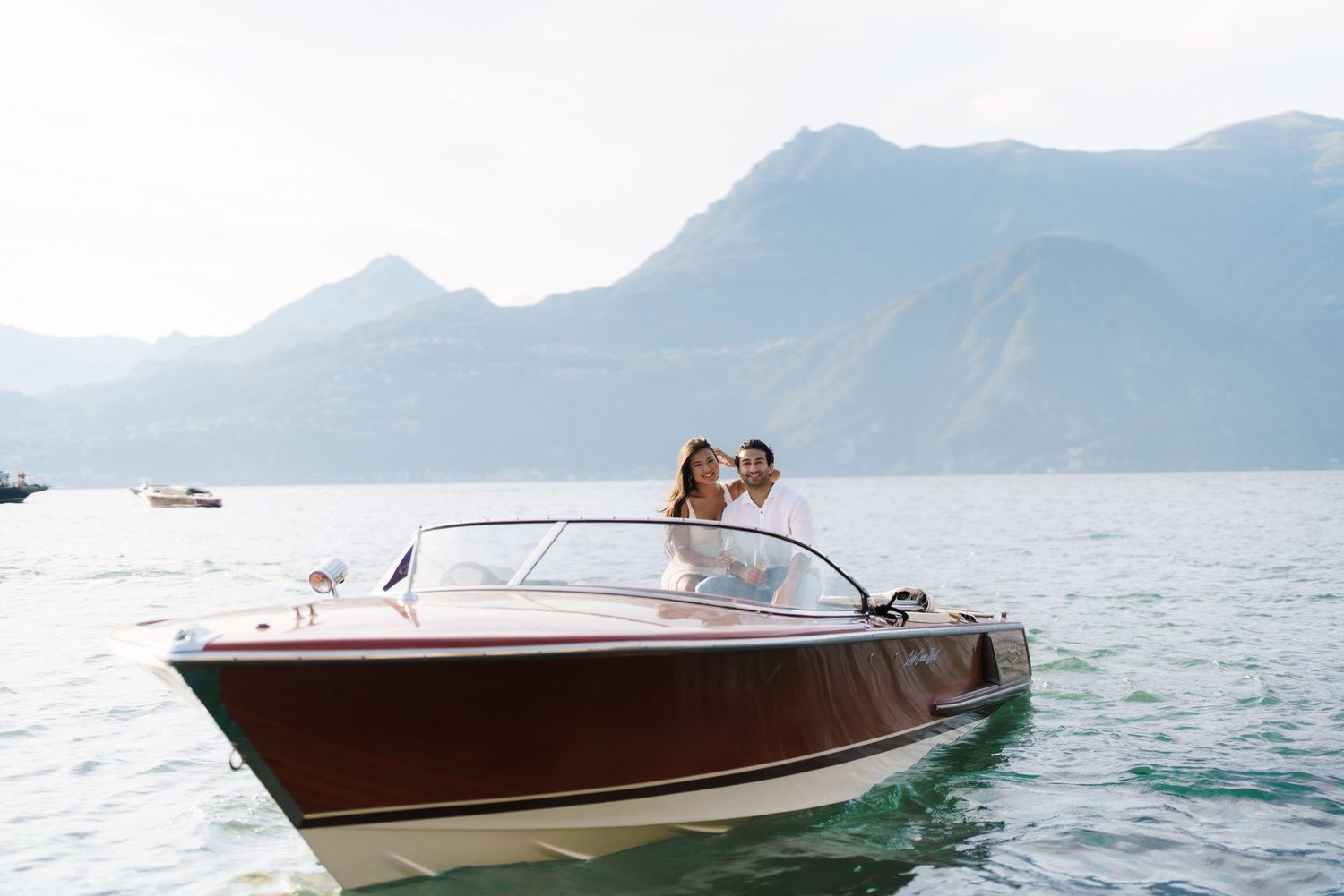Couple embracing on a classic wooden speedboat on a serene alpine lake with misty mountains in the background.