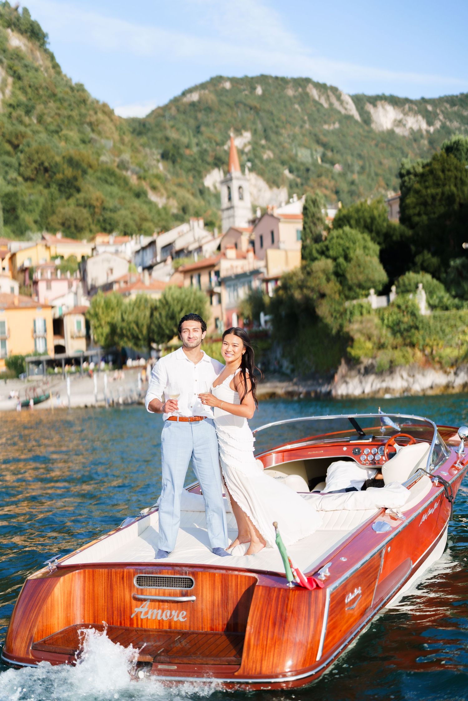 Couple standing on a classic wooden Riva boat named Amore on Lake Como, Italy, with colorful village backdrop.