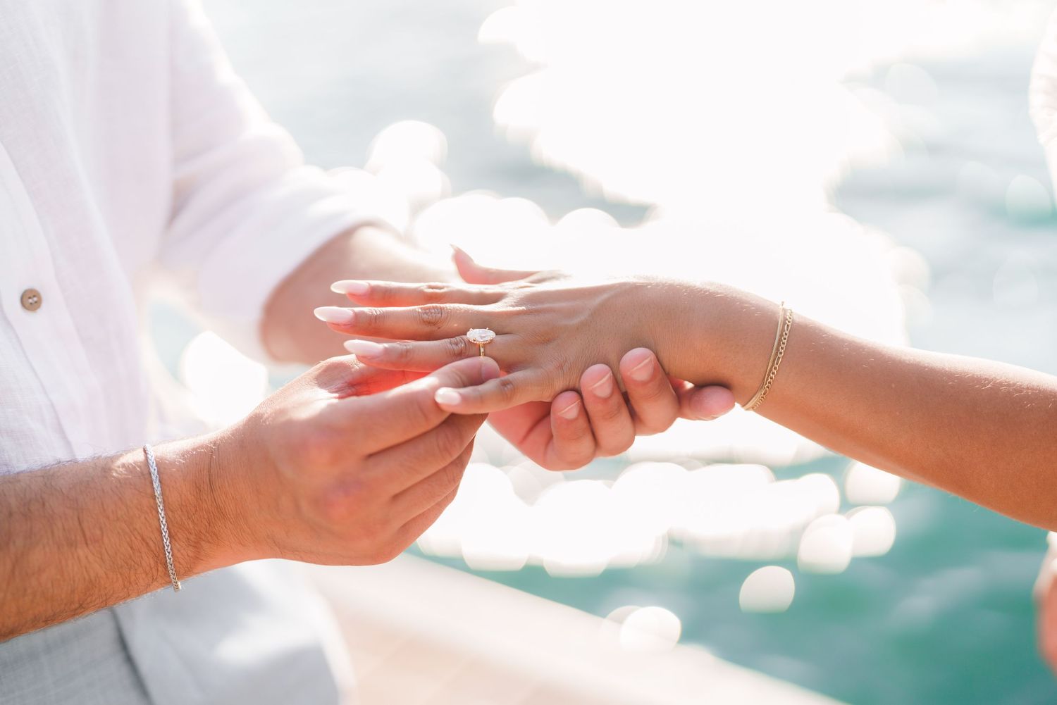Close-up of a man placing a wedding ring on a woman's finger outdoors near sparkling water.