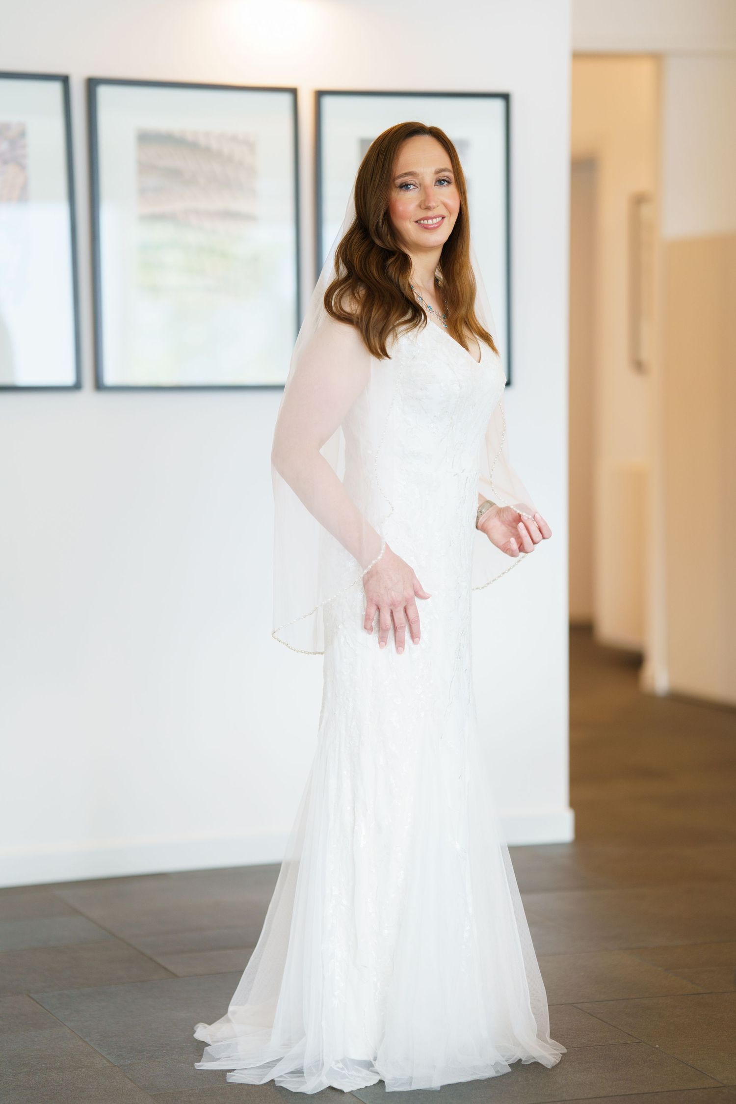 Smiling bride in elegant white lace long-sleeve wedding gown with sheer overlay, posing in a bright modern room.