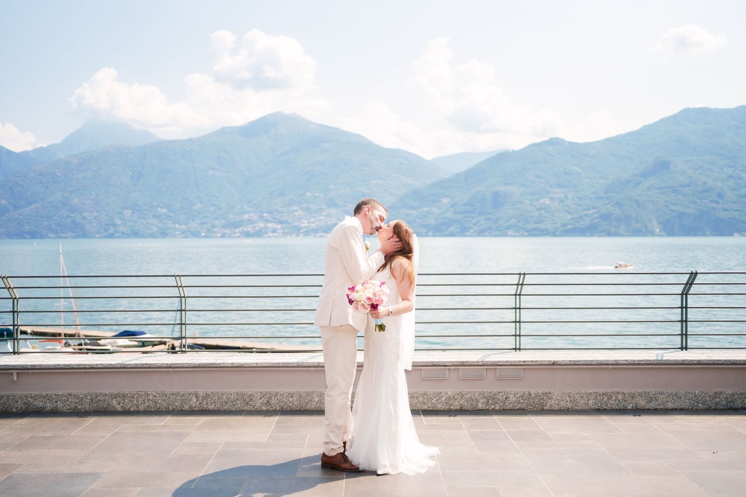 Bride and groom share a romantic kiss on a lakeside terrace with stunning mountain views in the background.