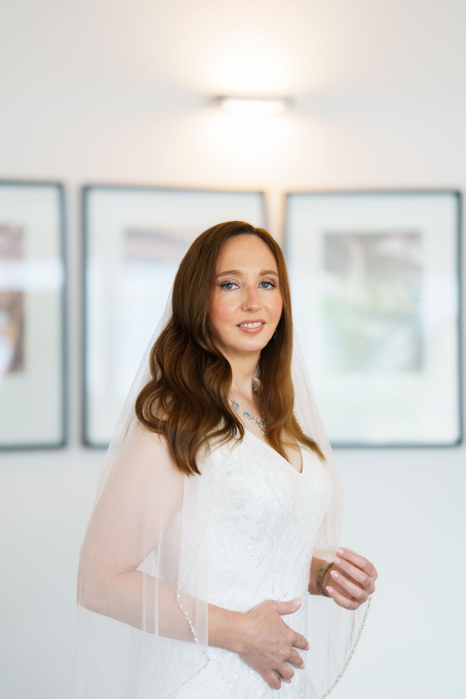 Elegant bride in white lace gown and veil smiling softly, wearing a diamond necklace in a bright modern room.
