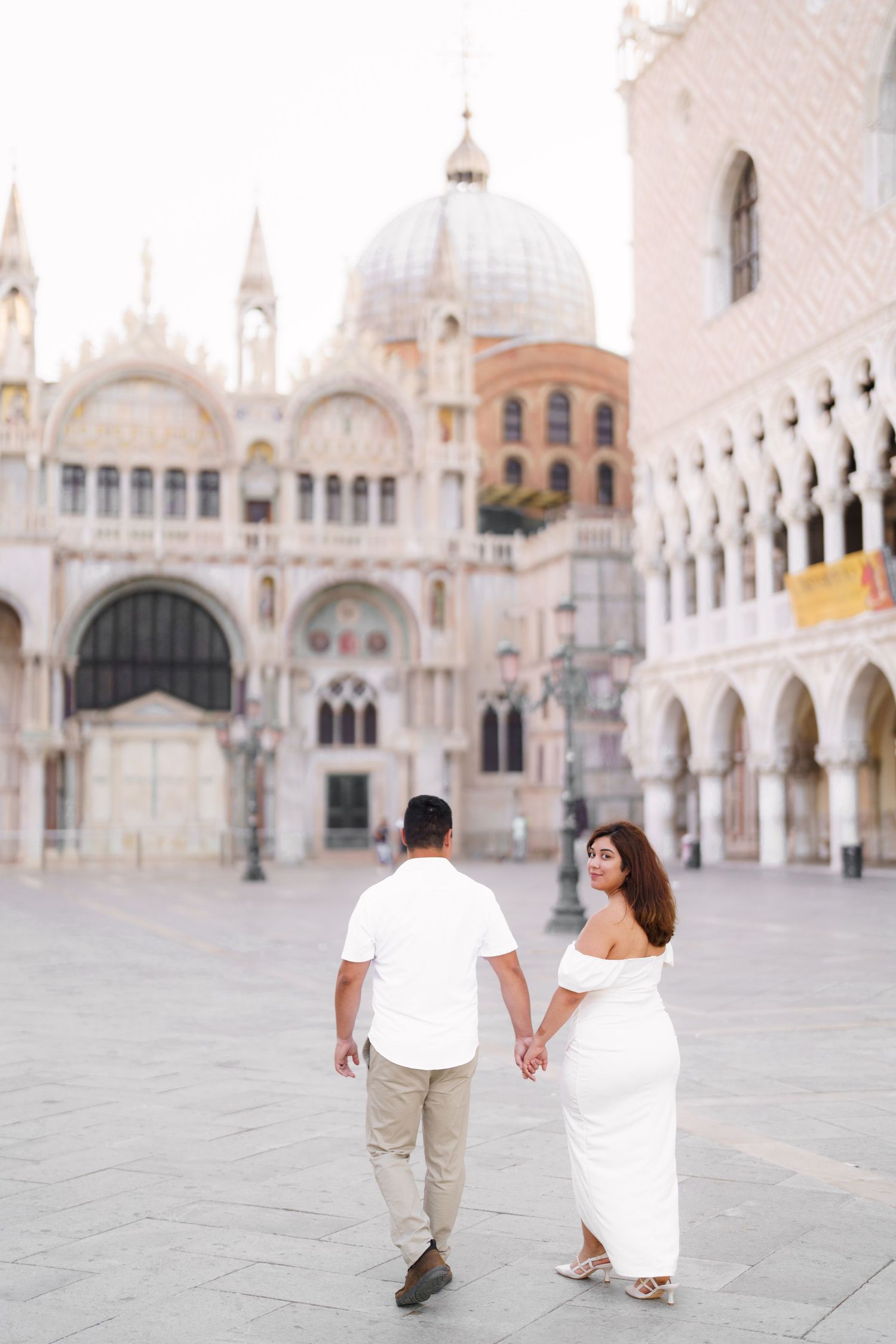 Romantic couple strolling hand in hand across empty St. Mark's Square with Venetian architecture behind them.