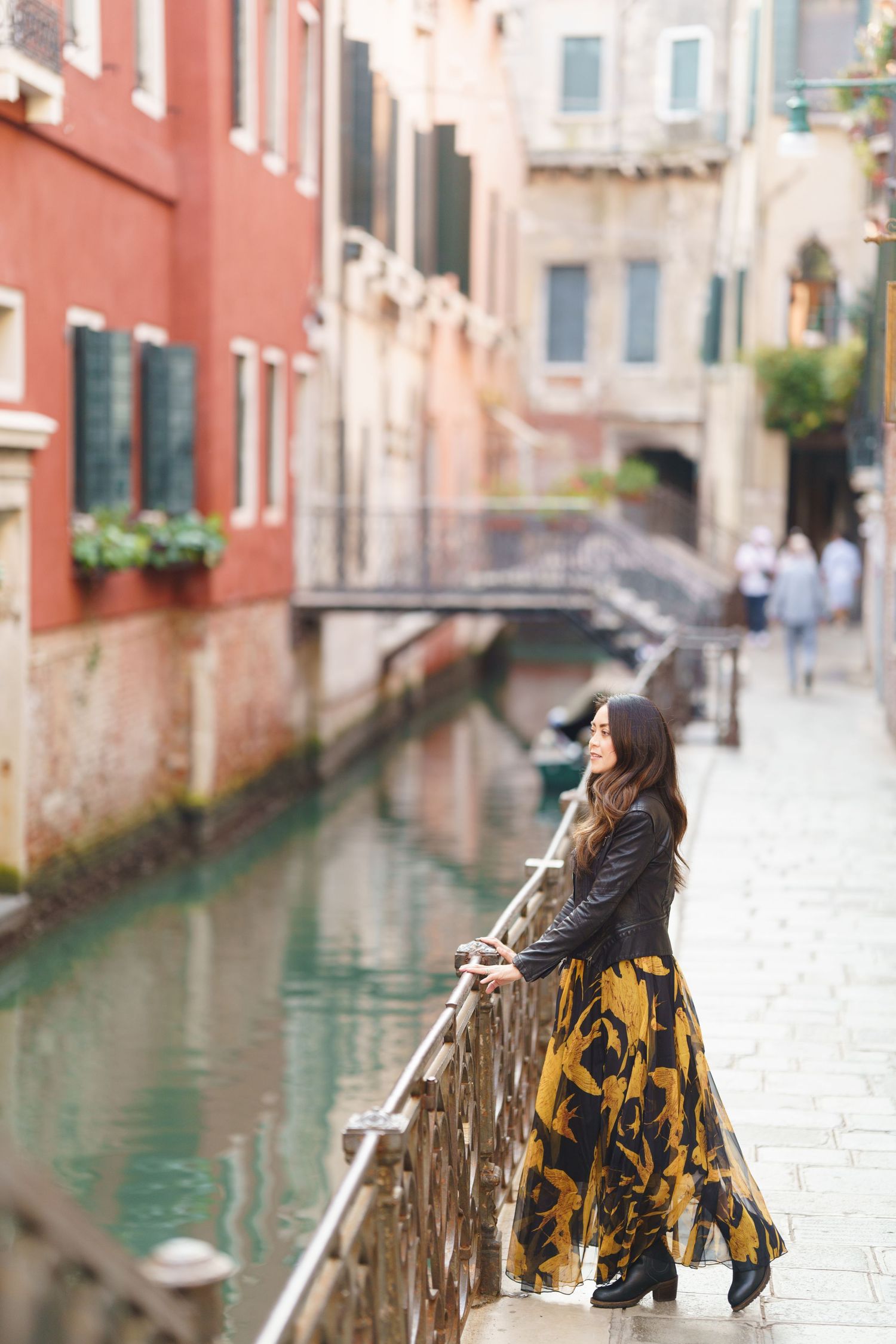 A person in a yellow skirt leans against a railing along a scenic Venice canal with pink buildings in the background.