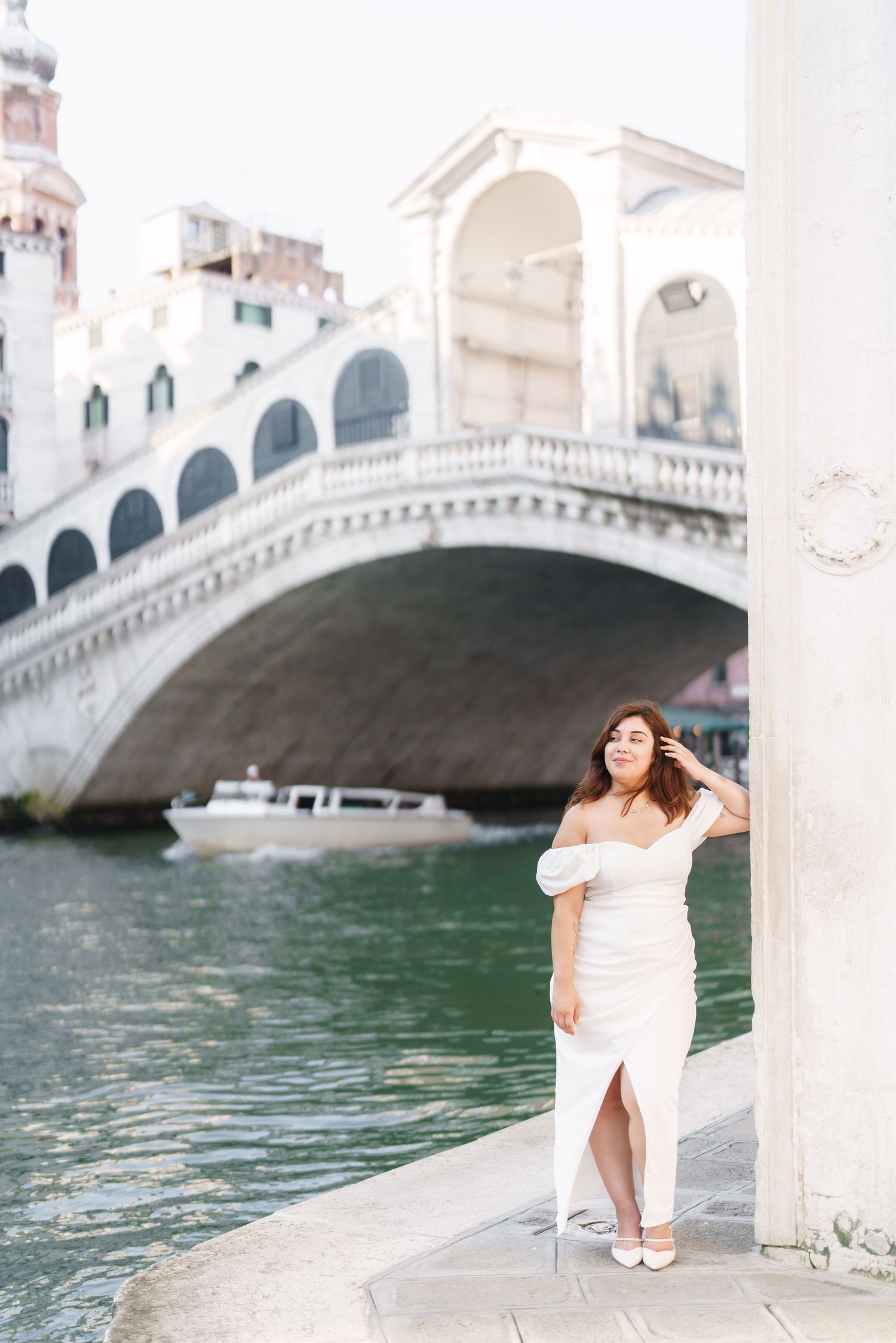 A figure in a white dress poses gracefully by a Venetian canal with the Rialto Bridge in the background.
