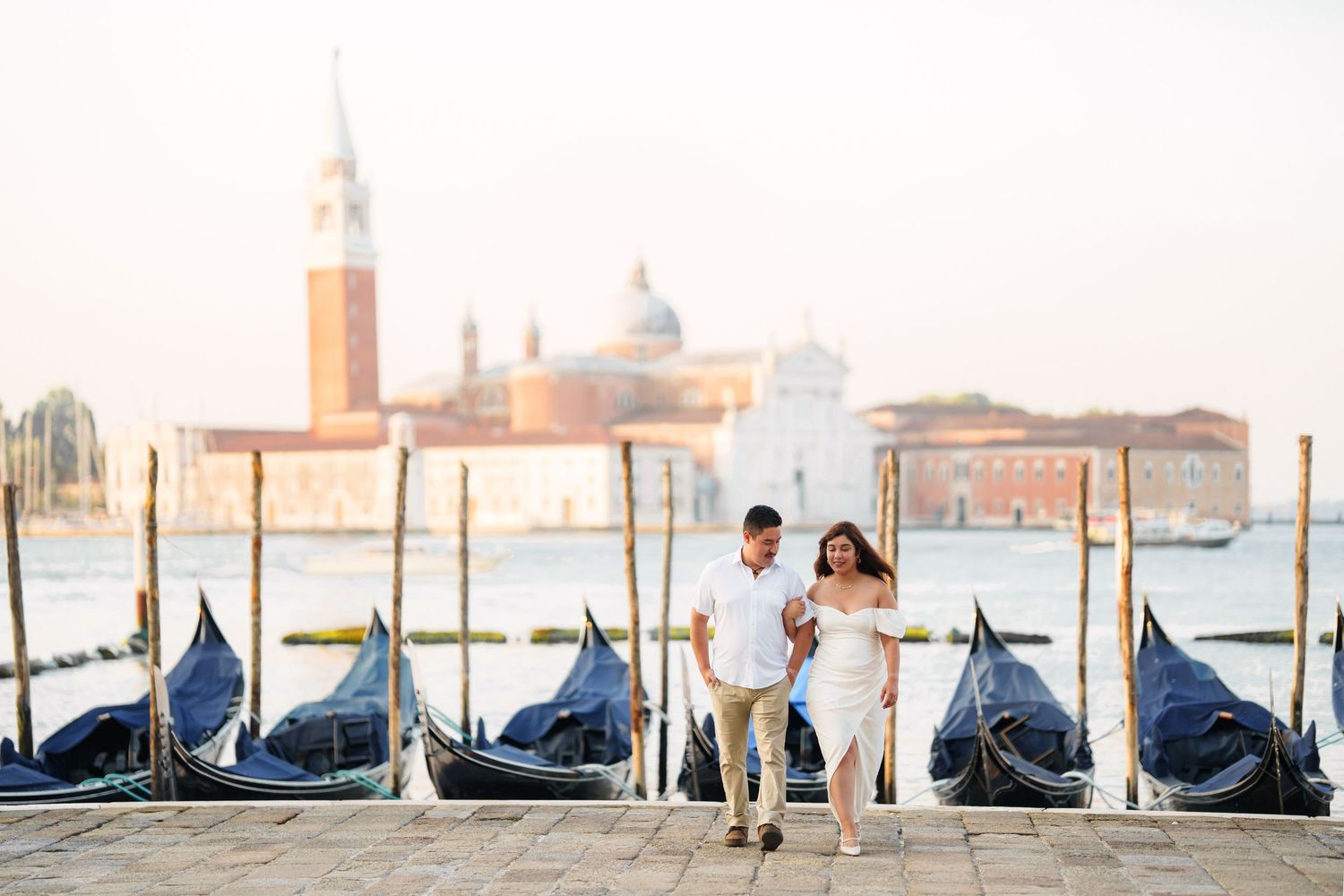 Romantic couple stands on Venice pier with iconic gondolas and San Giorgio Maggiore church in the background at sunset.