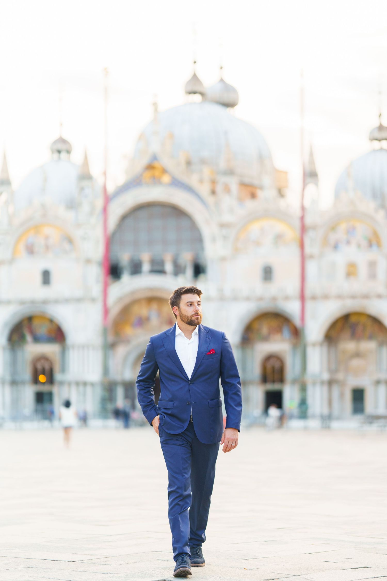 A person in a navy blue suit walks across St. Mark's Square in Venice with the basilica's ornate facade in the background.