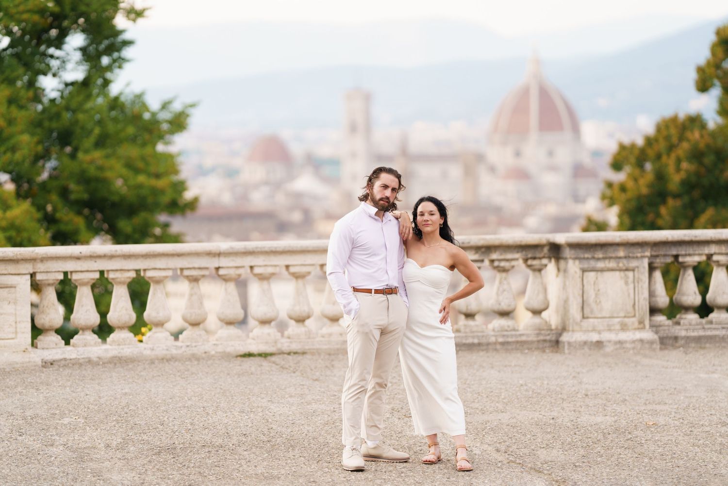 A couple in white outfits poses for romantic photos on a scenic overlook with Florence, Italy in the background.