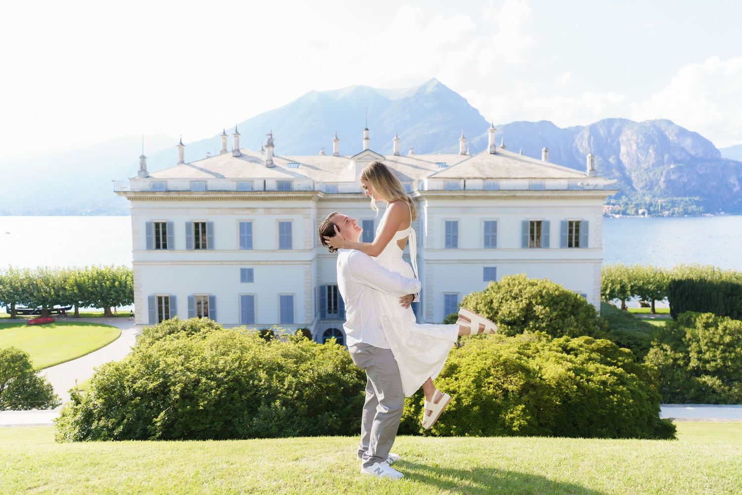 A romantic moment captured on a grand estate lawn overlooking Lake Como with mountains and a white villa in the background.