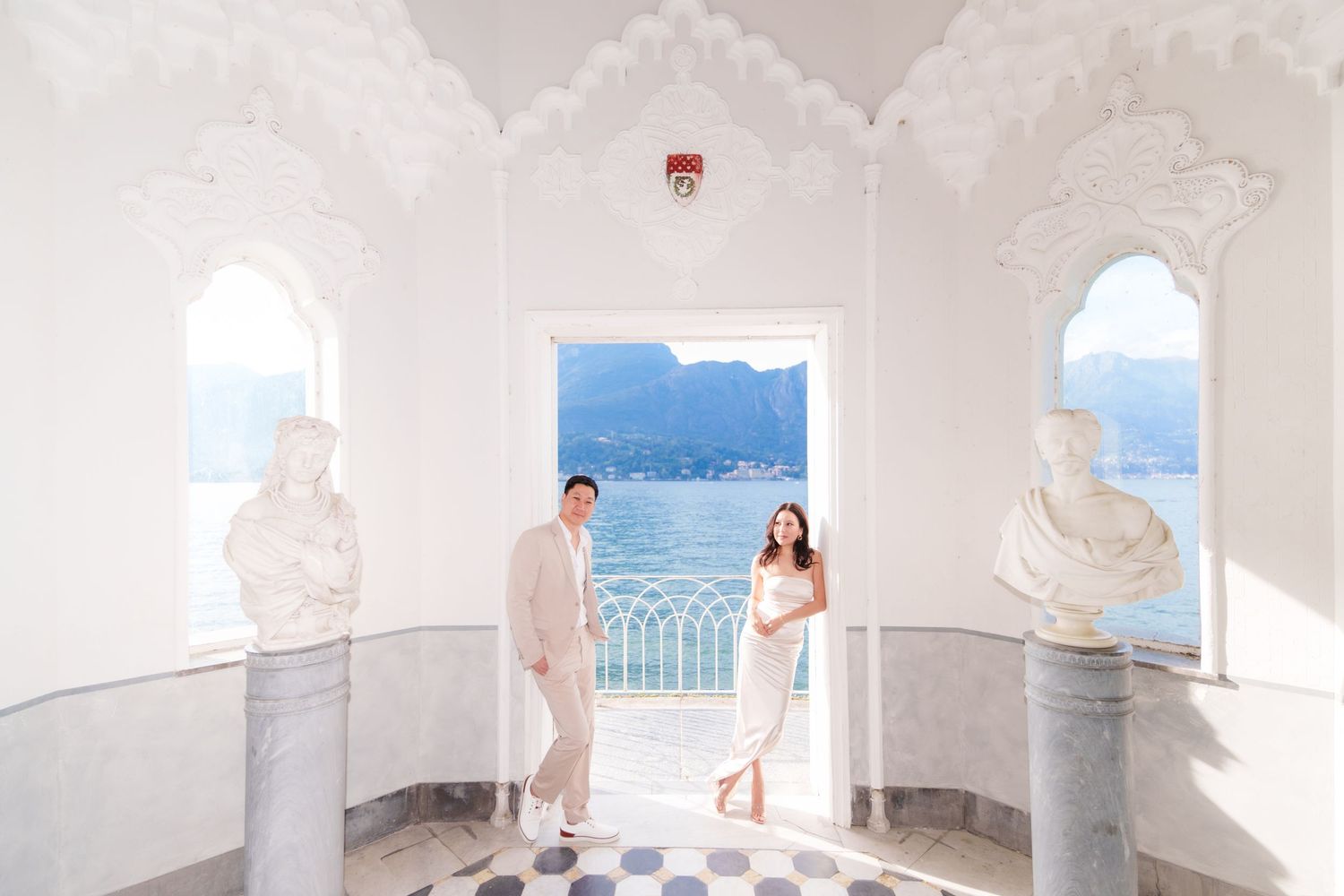 A couple stands in a white Mediterranean archway overlooking a scenic coastal view with blue water and mountains.