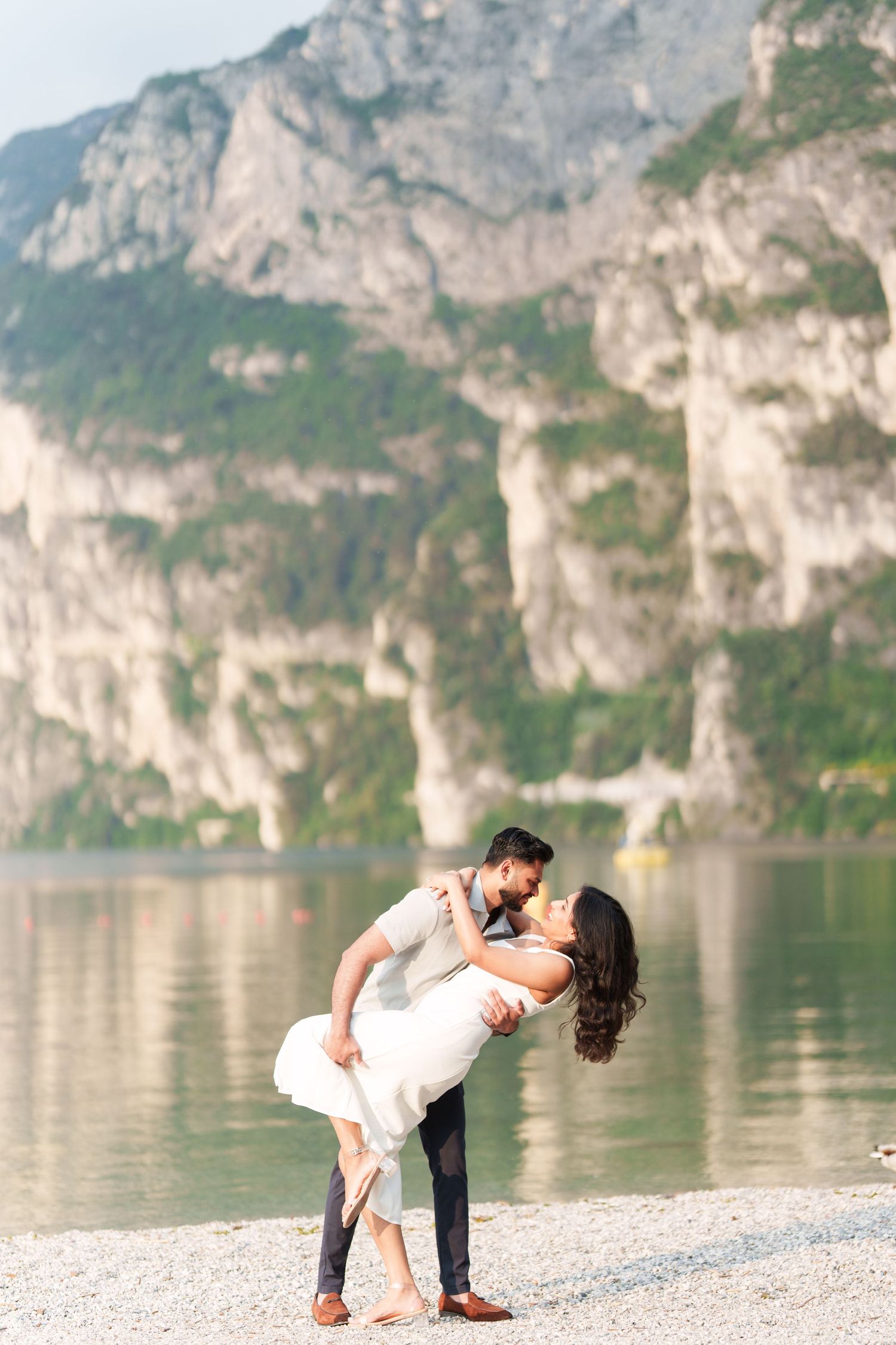 A romantic couple shares a tender dip pose on a scenic rocky beach with dramatic mountain cliffs and lake in the background.