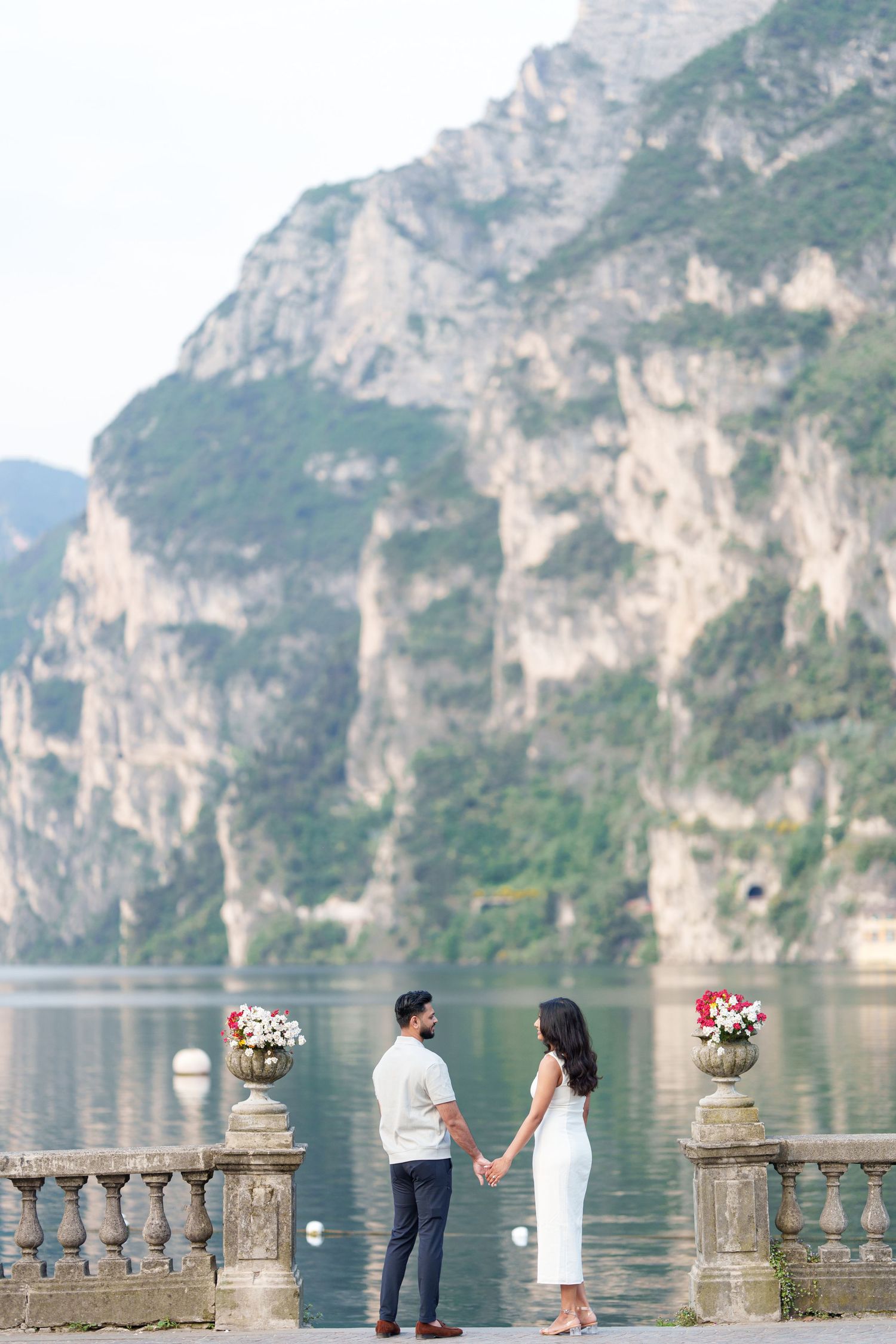A couple stands by a stone balustrade overlooking a scenic mountain lake surrounded by towering limestone cliffs.
