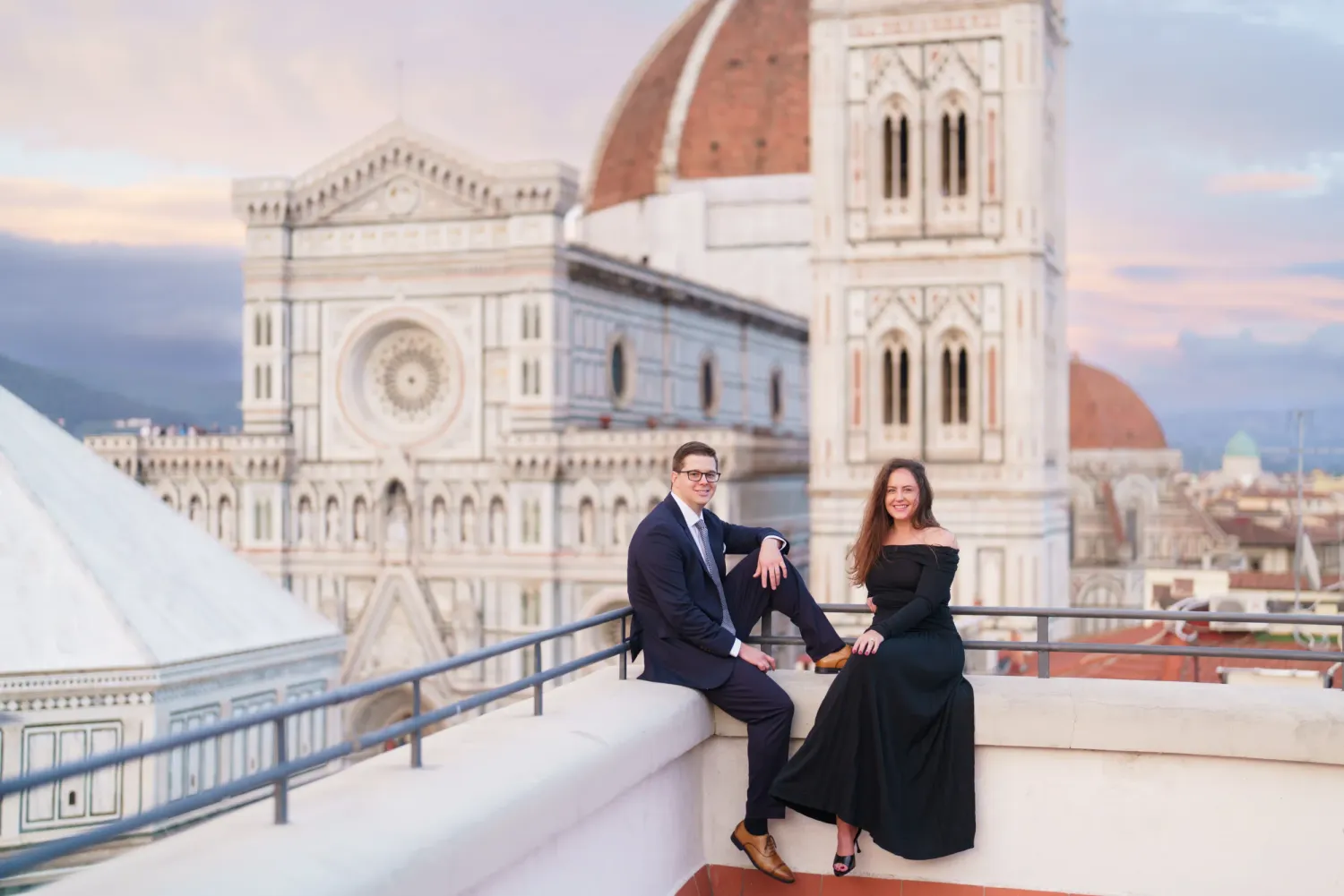 Couple poses on rooftop terrace overlooking Florence Cathedral and historic cityscape during sunset in Italy.