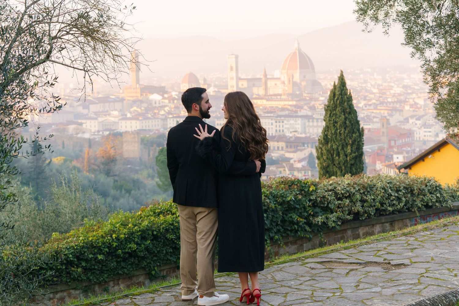 A couple stands together overlooking Florence, Italy at sunset with the iconic Duomo cathedral visible in the distance.