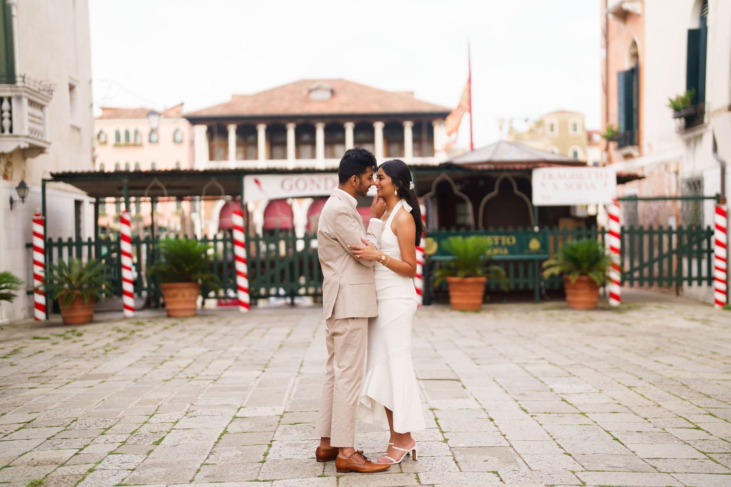 A couple in light-colored outfits walks hand in hand through a charming European cobblestone plaza.