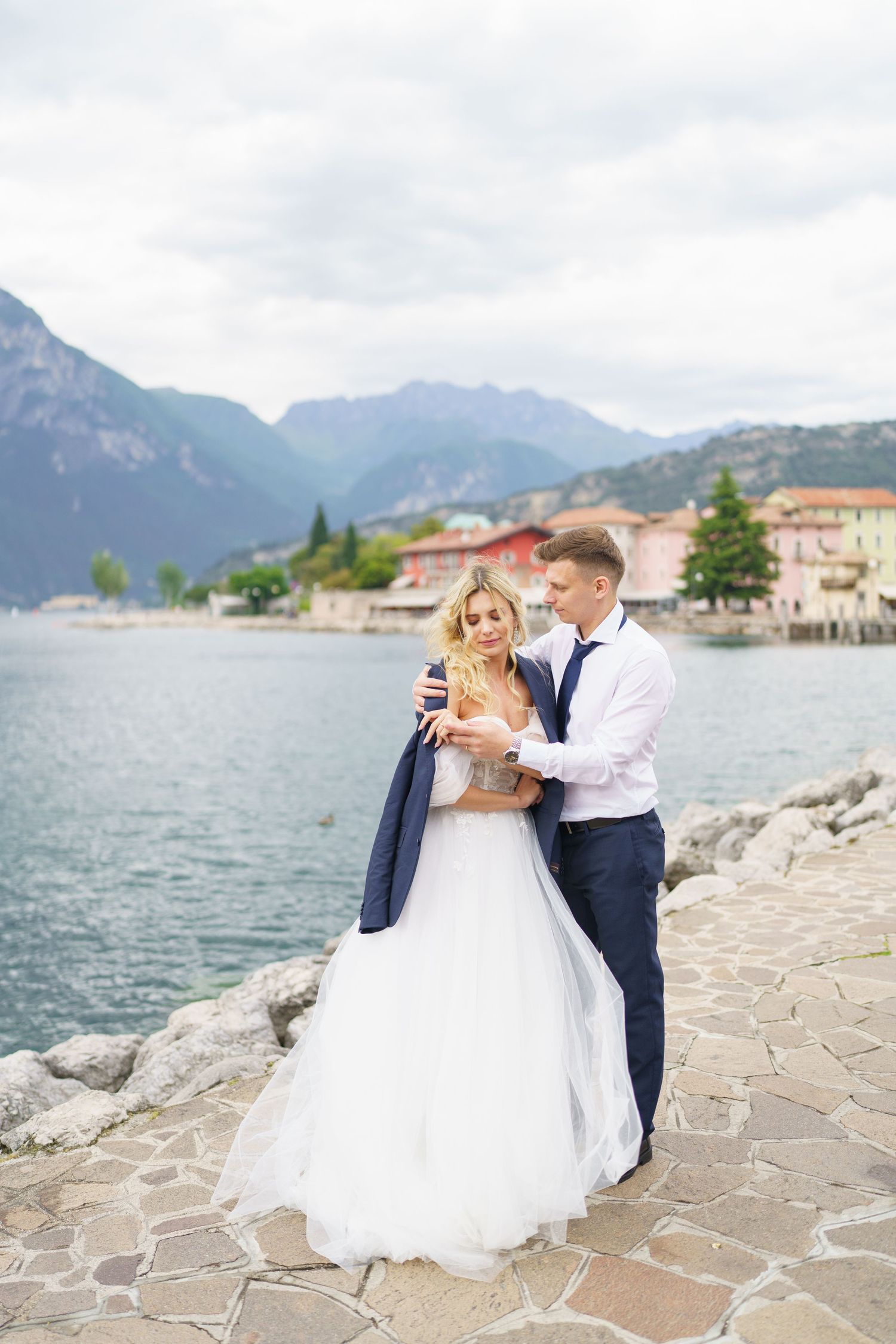 A couple in wedding attire embrace on a stone walkway by a lake with mountains and Italian buildings in the background.
