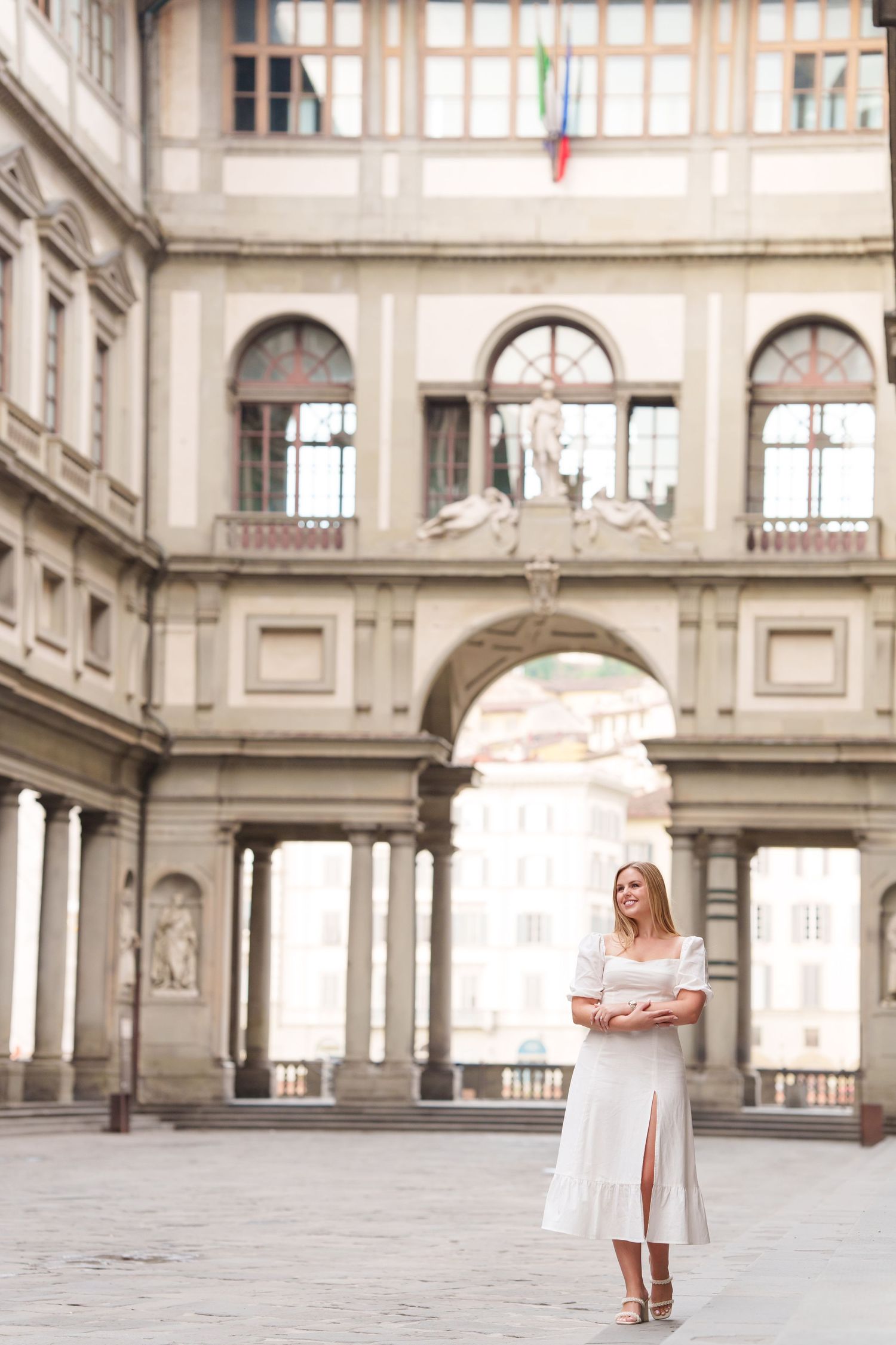 A figure in a flowing white dress poses gracefully in front of ornate Italian Renaissance architecture at the Uffizi Gallery.