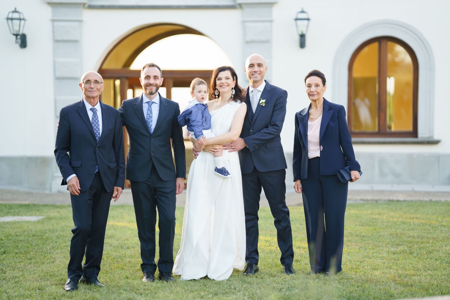 Wedding party gathered outside elegant white building with arched architecture.