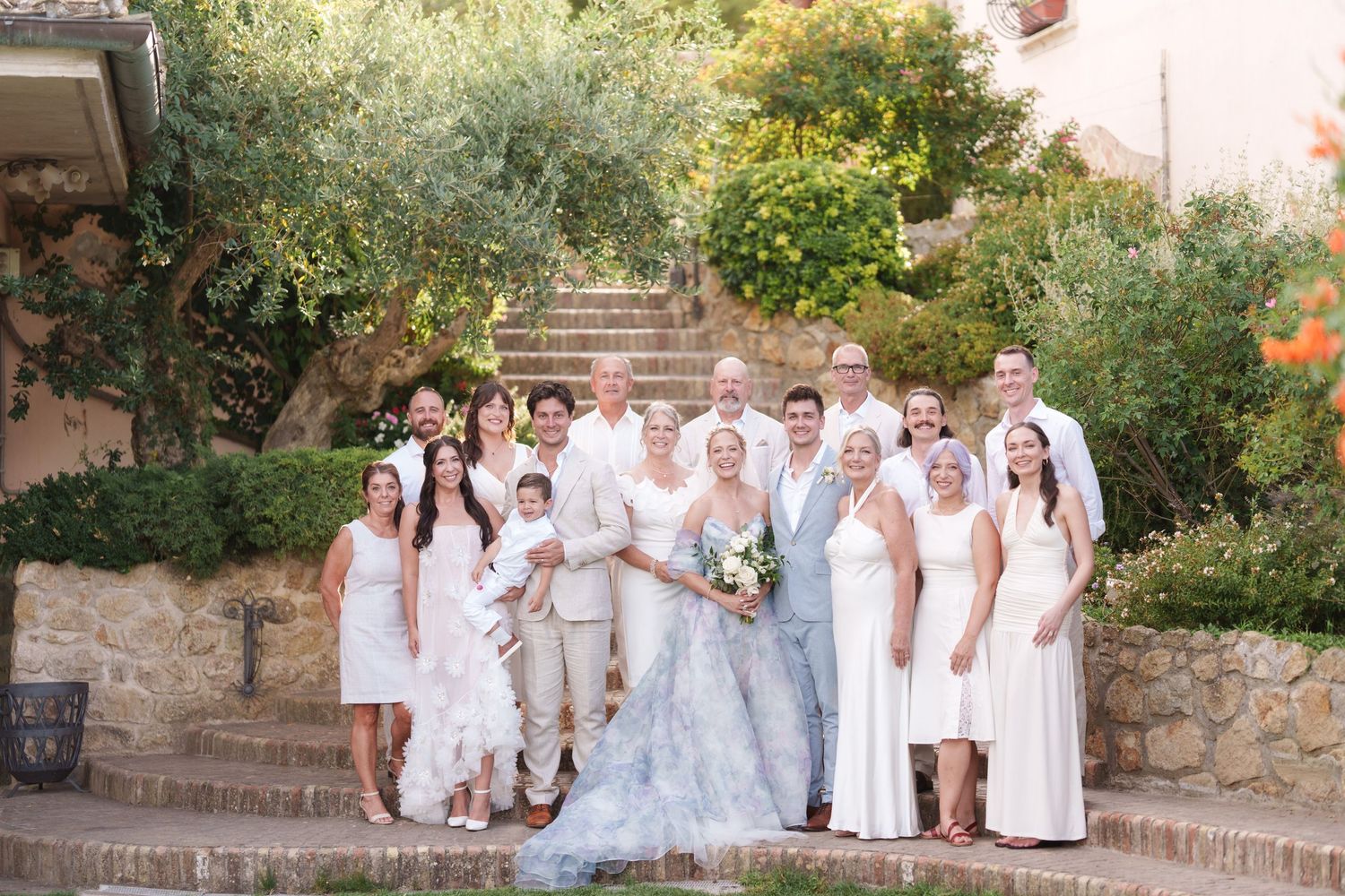 Wedding party poses on stone steps surrounded by lush greenery, bride in blue gown with white-dressed guests.