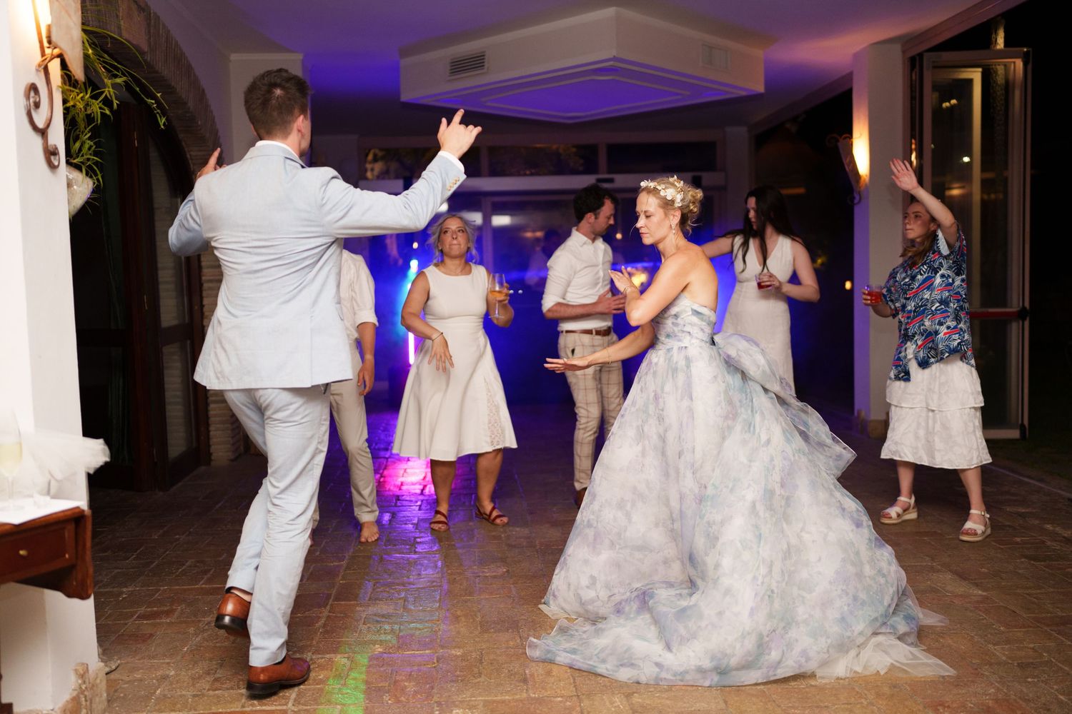 Bride and groom dancing joyfully at wedding reception with guests celebrating in elegant venue with blue lighting.