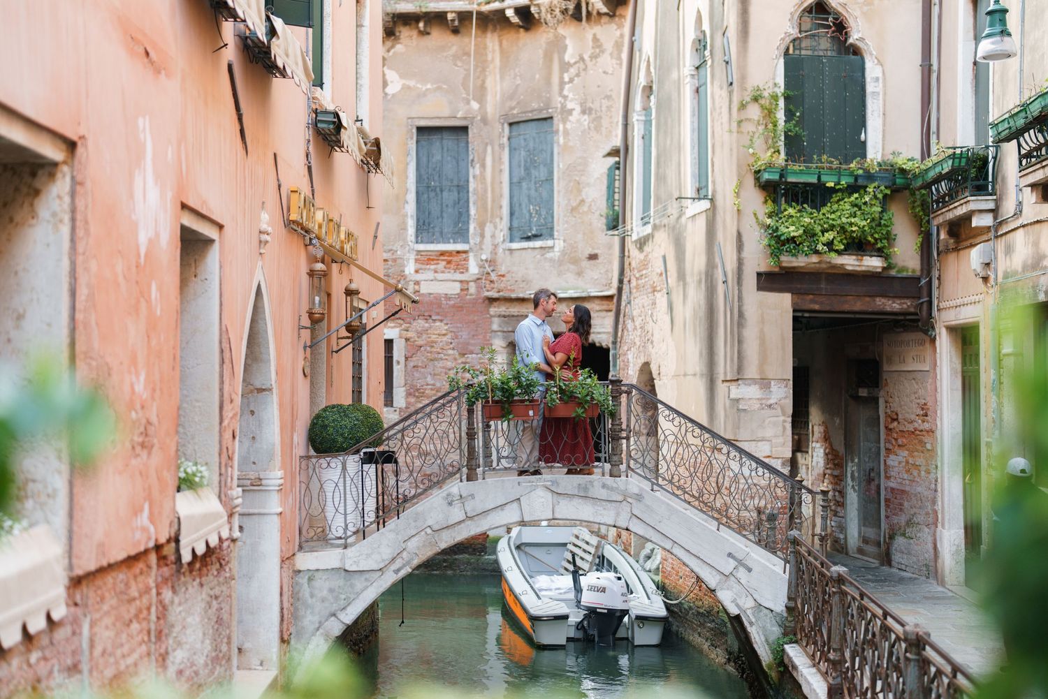 Couple embracing on a stone bridge over a Venice canal with colorful historic buildings and moored boats.
