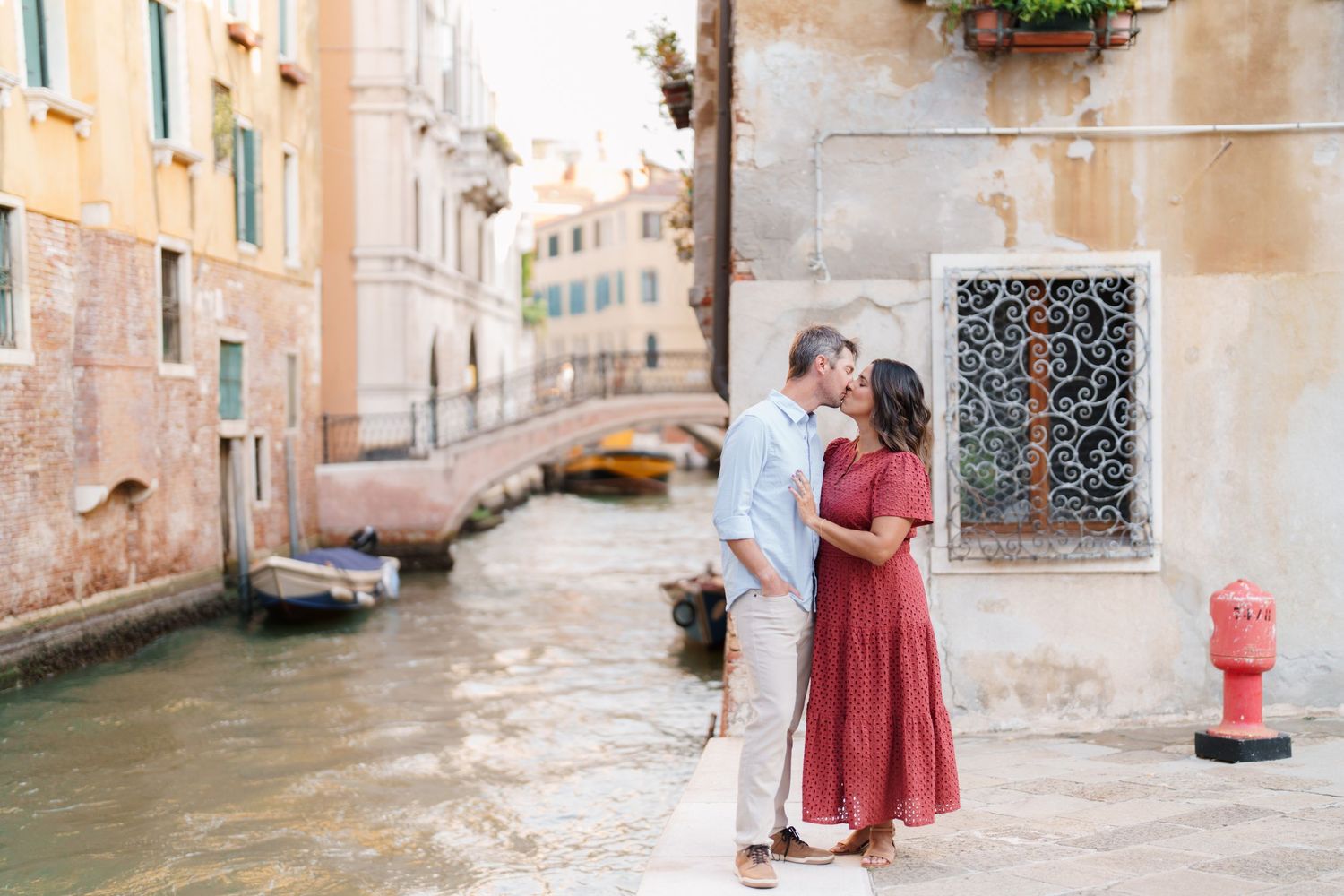 Couple kissing romantically on a Venice canal walkway, surrounded by colorful historic buildings and boats.