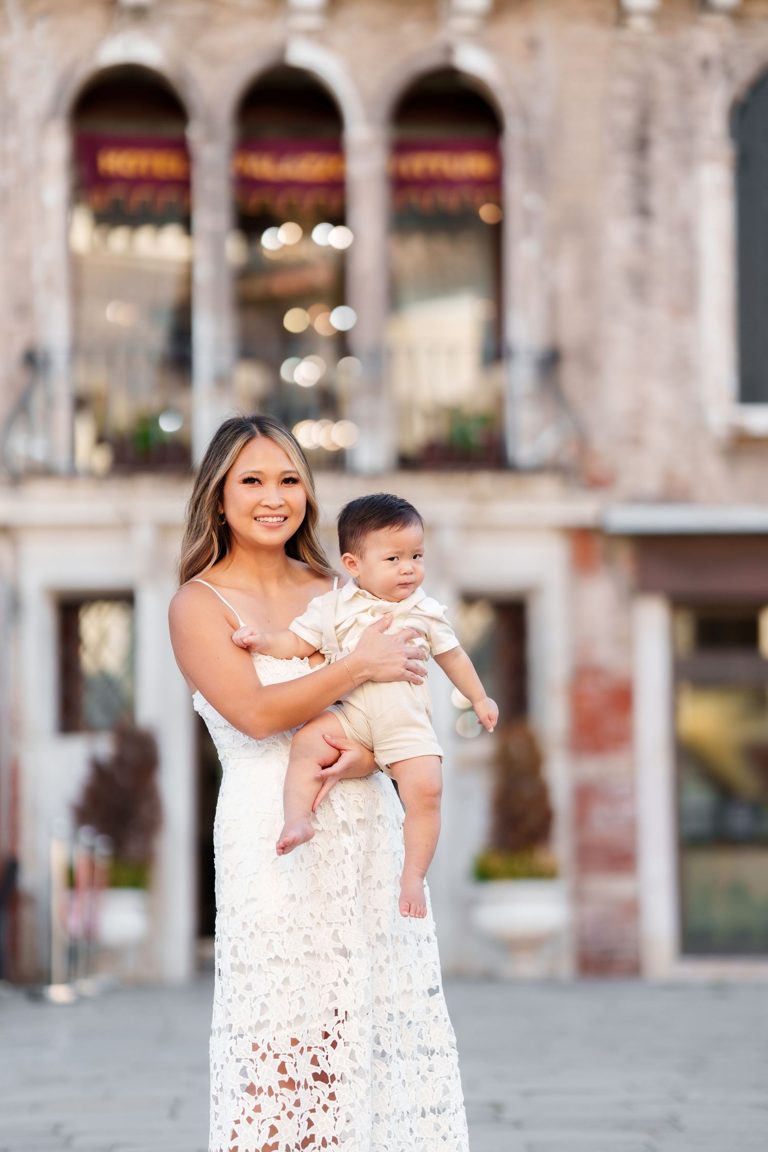 A smiling woman in a white lace dress holds her baby boy in a sunny European plaza.