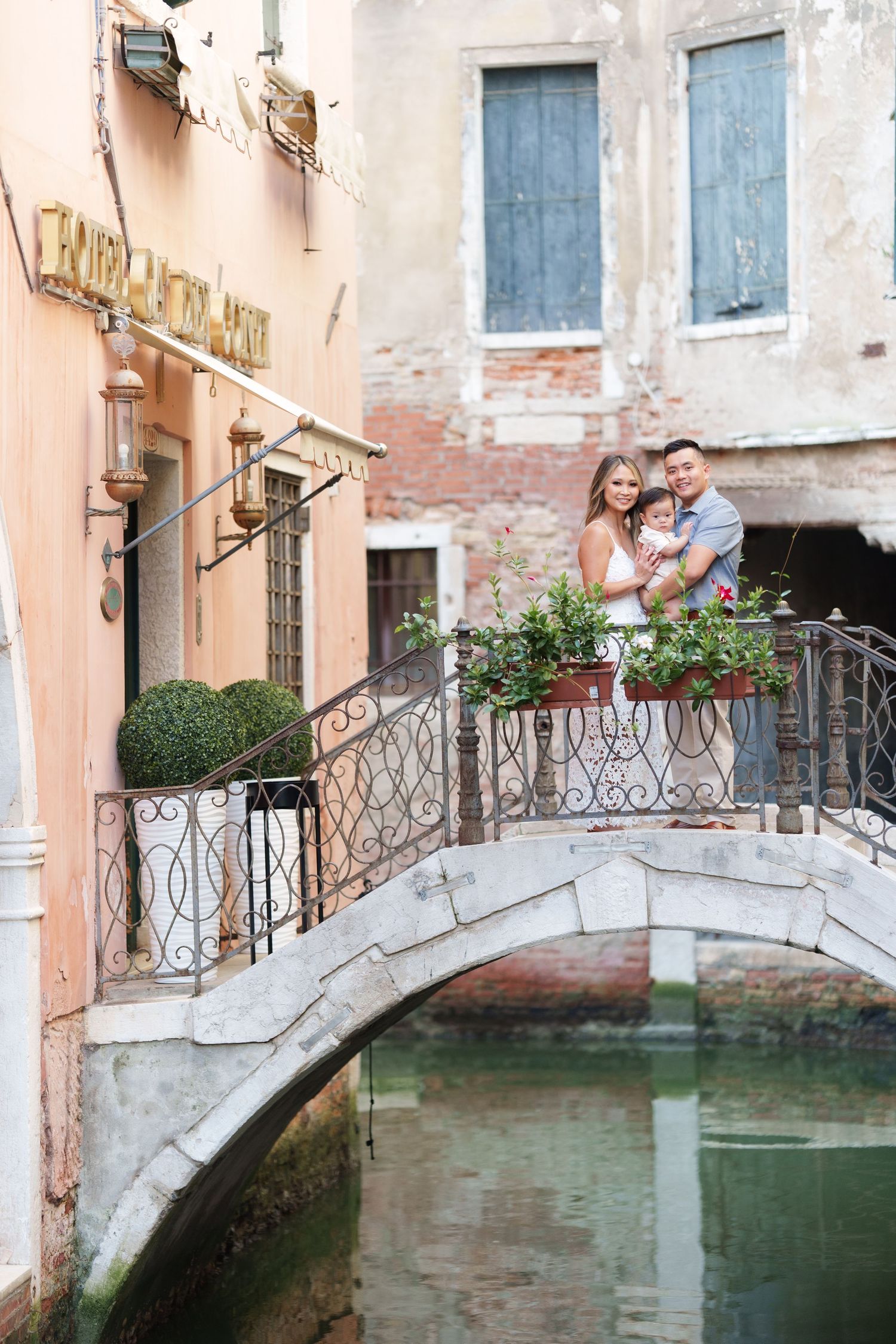 A happy family poses on a charming stone bridge over a Venice canal, surrounded by colorful flowers and historic buildings.